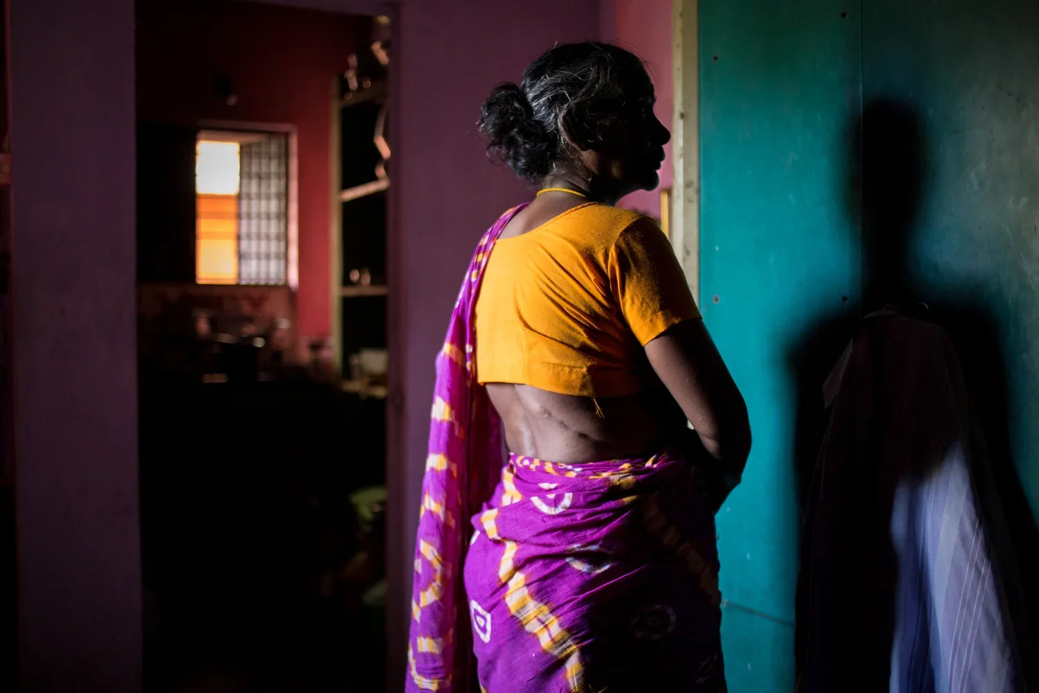 Mulher de cabelo cacheado, vestindo blusa amarelo e saia roxa com padrão, de costas, encostada na parede azul, em ambiente interno com janelas ao fundo.