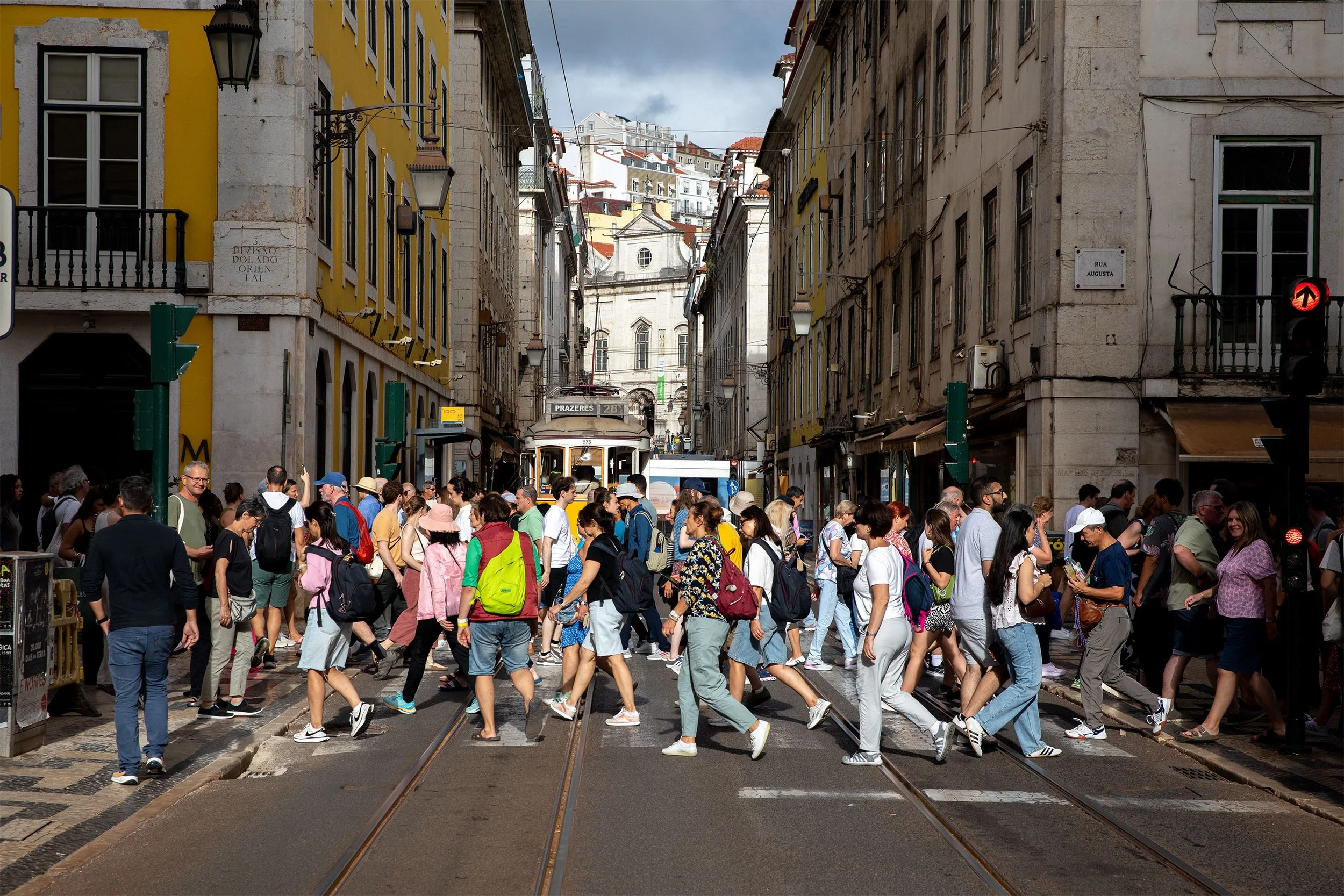 Muitos pedestres atravessando a rua em uma cidade, com um bonde ao fundo e prédios históricos nas laterais.