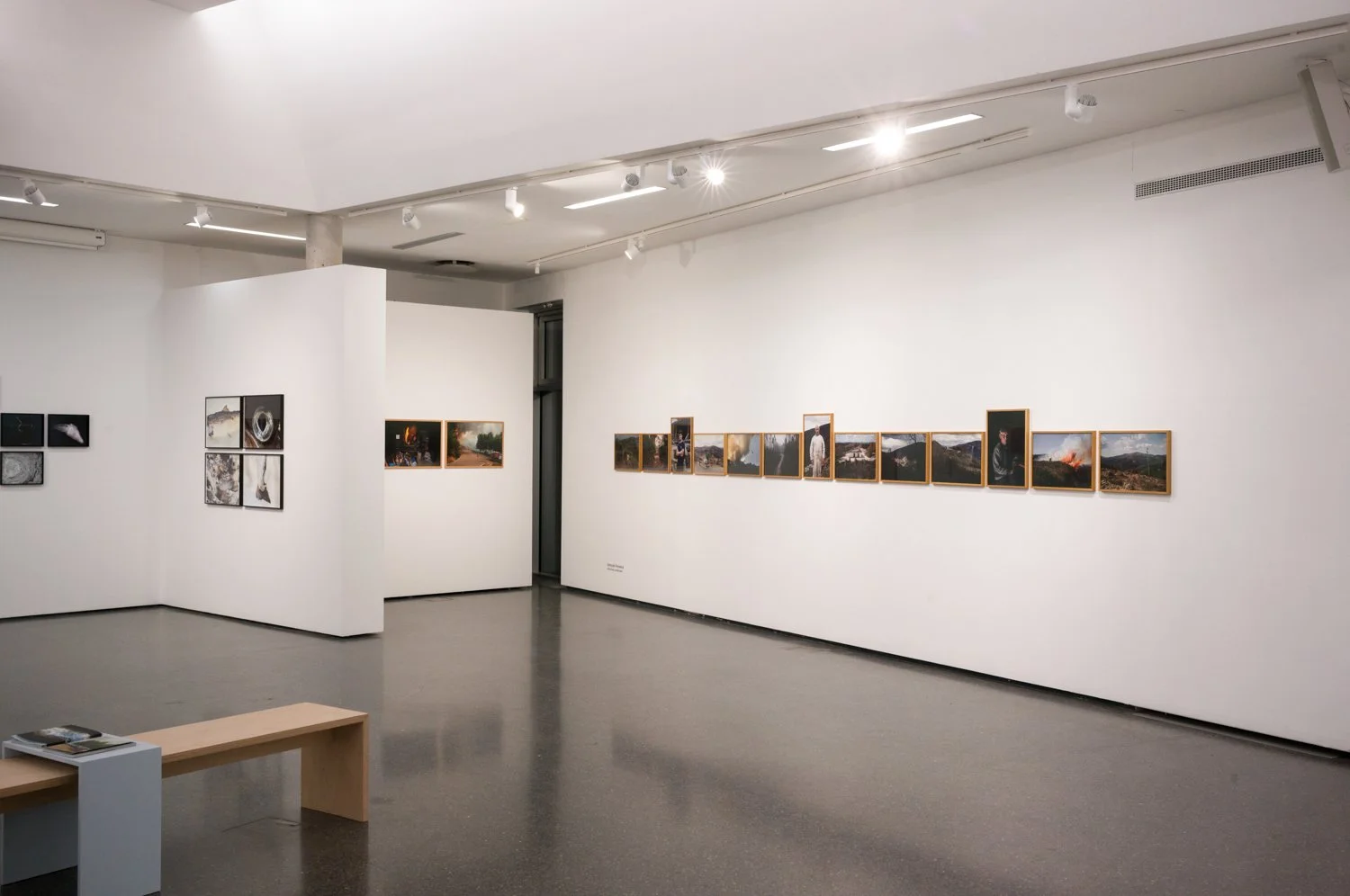 Sala de exposição de arte com várias fotografias em suas paredes brancas, com iluminação focada nas obras e um banco na frente.