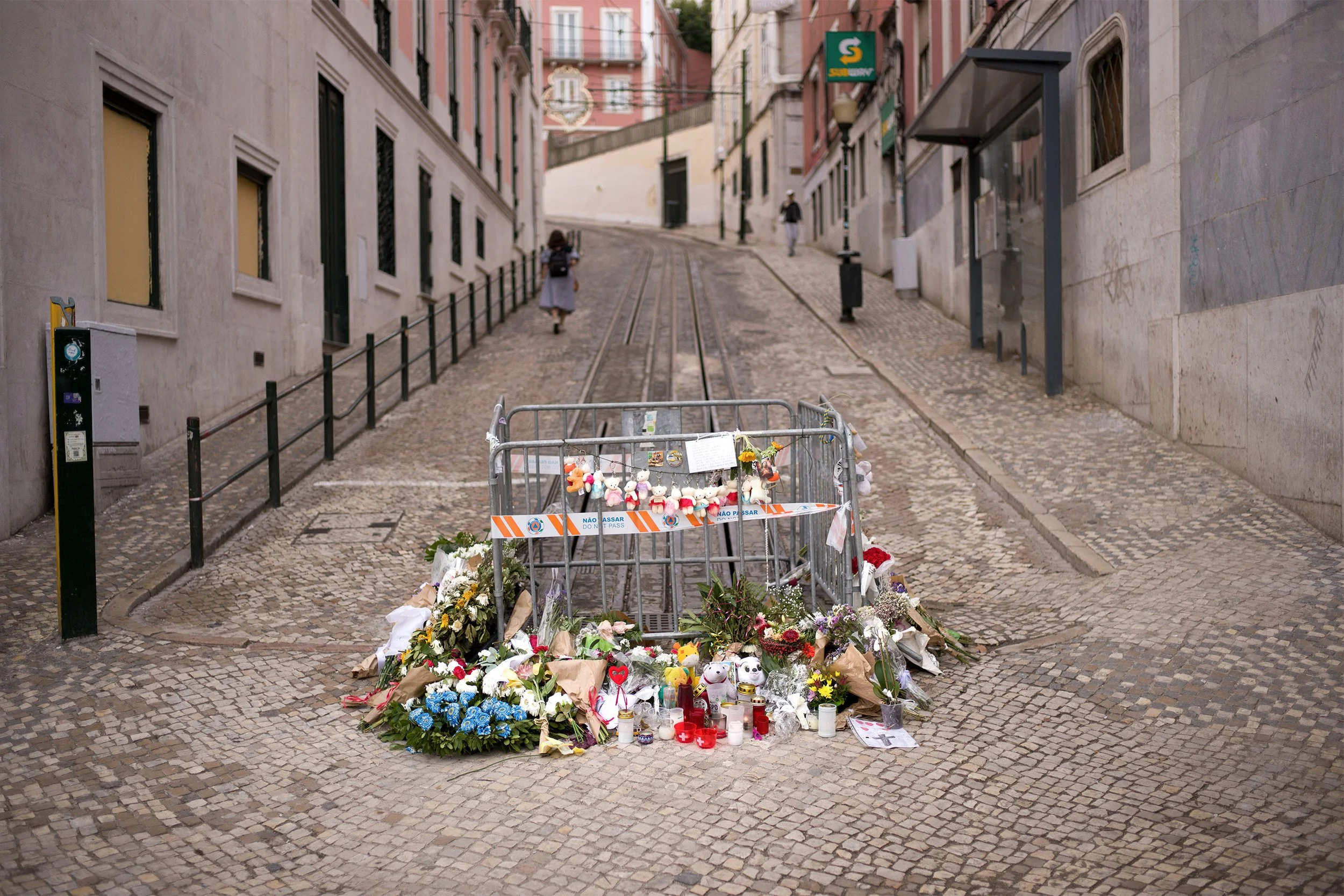 Procissão de flores e velas em homenagem na rua com chão de calcário, ao redor de uma cerca com aviso de não passar.