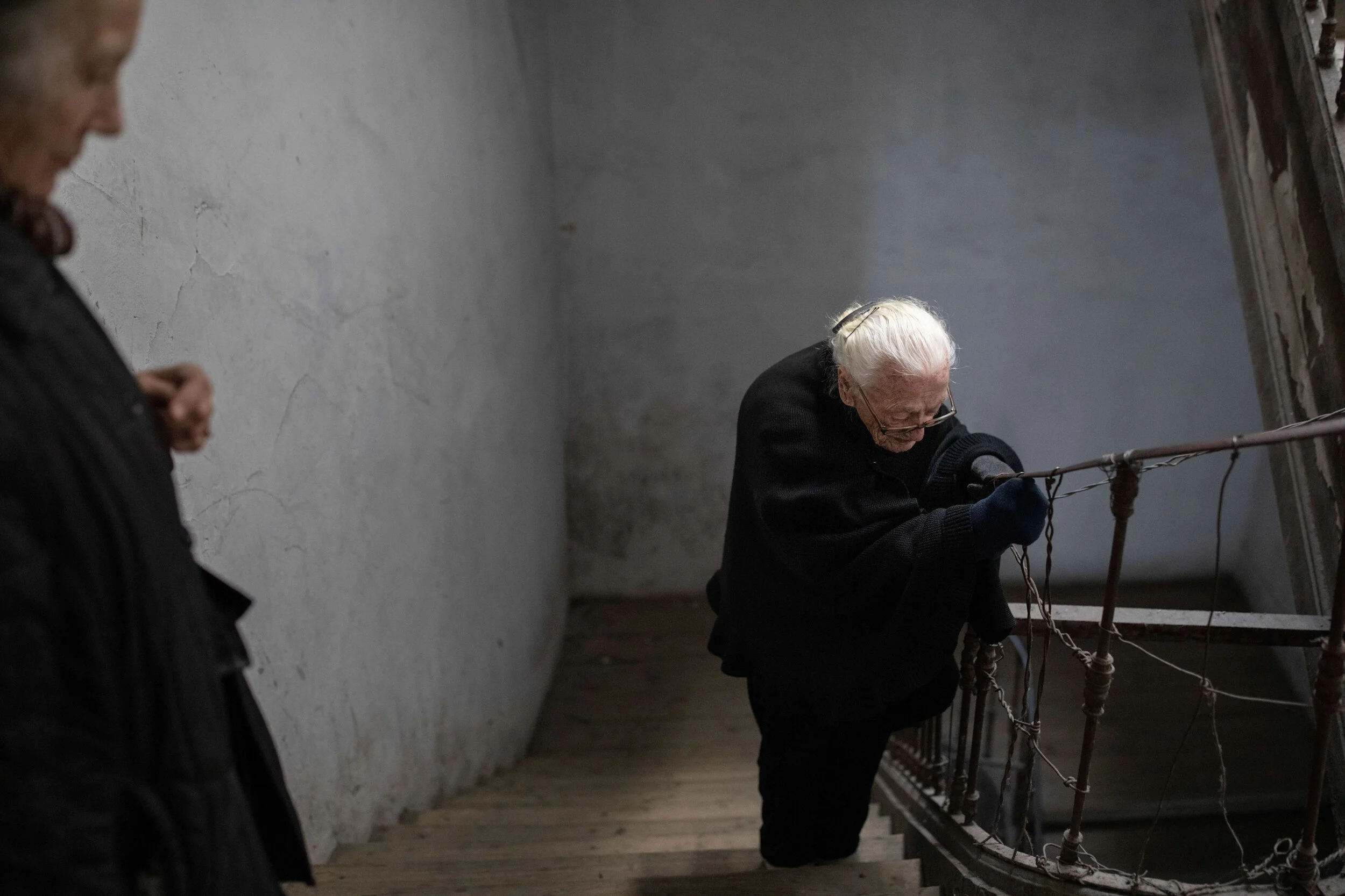 Duas pessoas idosas em uma escada de ferro com corrimão de corda, uma delas está inclinada e segurando na grade, enquanto a outra observa ao lado.