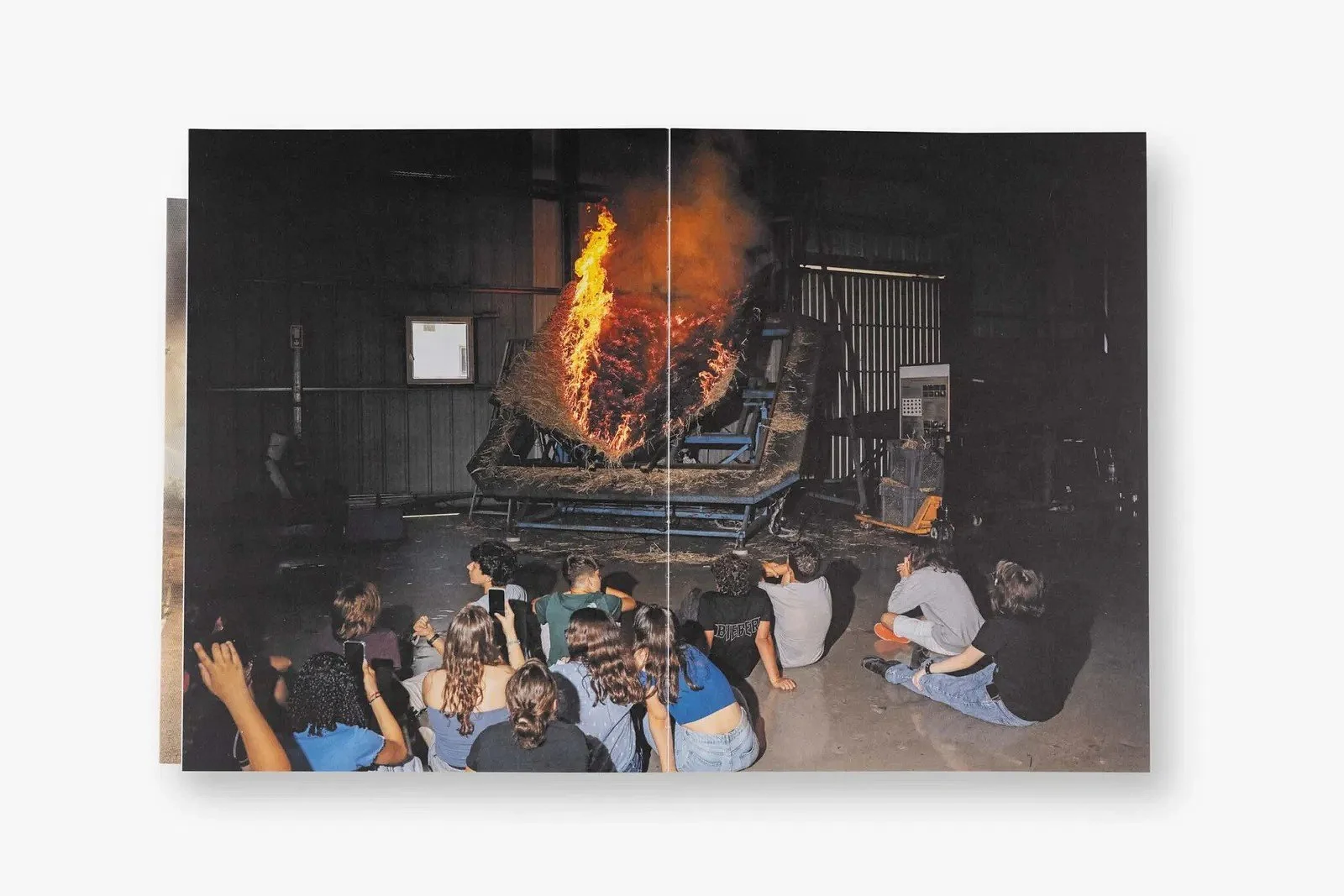 Grupo de pessoas assistindo a uma performance com uma cama inflável em chama, dentro de uma estrutura de galpão.