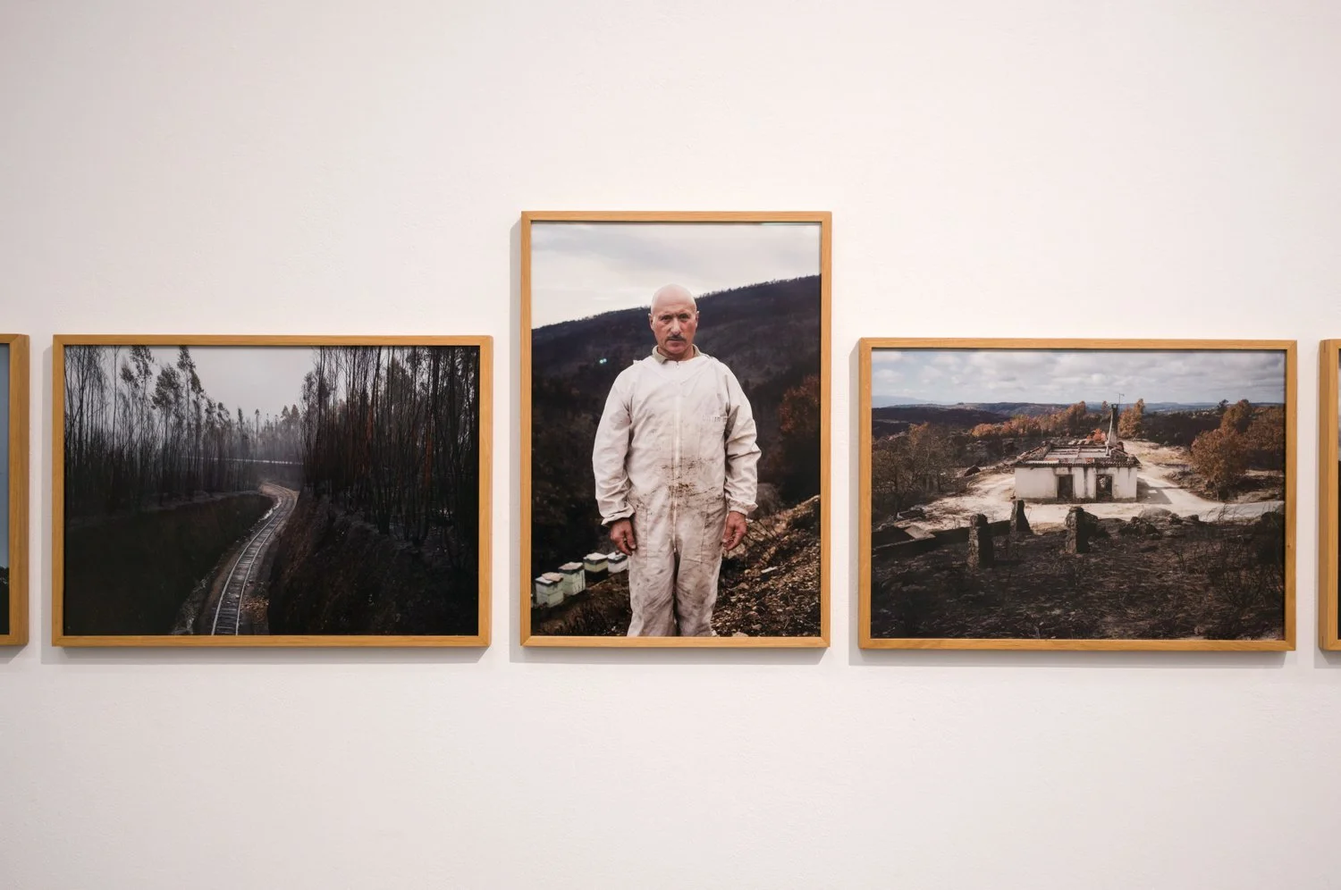 Fotografia de uma galeria de arte com três quadros emoldurados na parede. No centro, uma pessoa vestindo roupa branca, com aparência cansada, de cabeça rapada, posando diante de uma paisagem montanhosa.