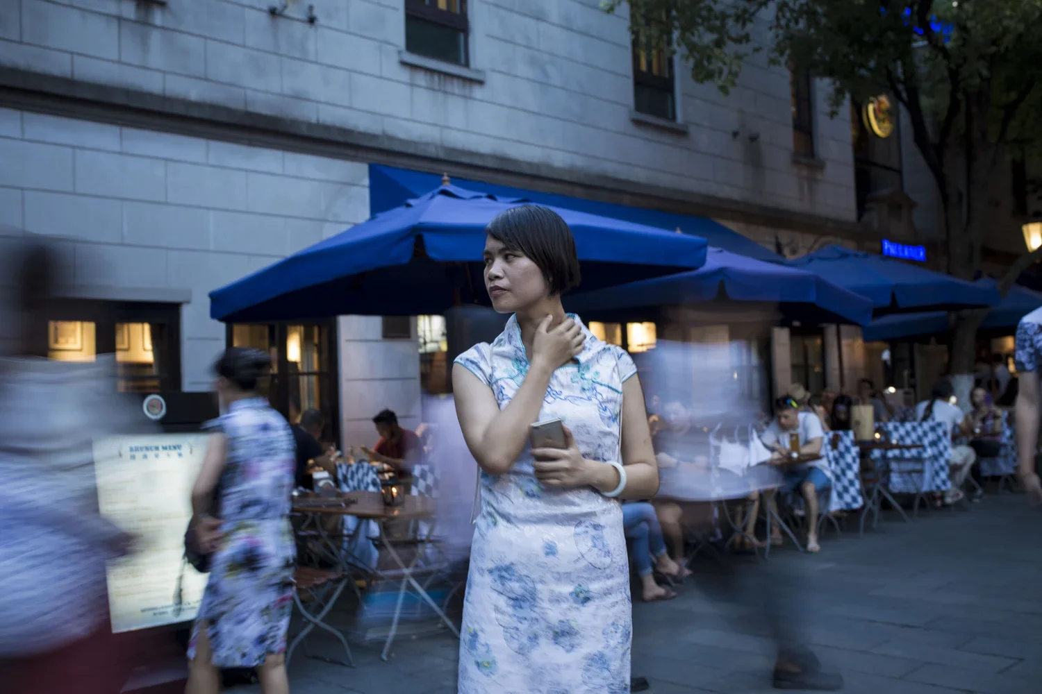 Mulher com cabelo curto usando vestido com estampa azul e branco, segurando um telefone, em rua com pessoas sentadas em mesas ao ar livre sob guarda-sóis azuis, no entardecer ou início da noite.