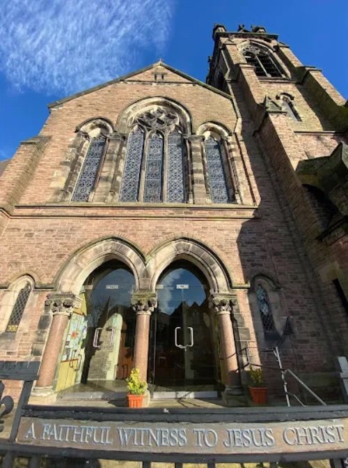 View of a tall stone church with arched stained glass windows, an entrance with glass doors, and a bell tower under a blue sky with wispy clouds. A metal gate and potted plants are in front, with an inscription on the gate reading 'A Faithful Witness to Jesus Christ.'