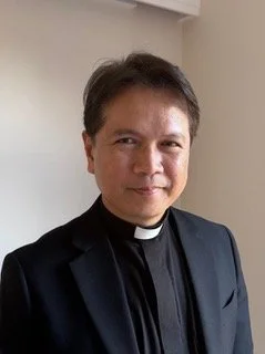 A man dressed in clerical clothing, including a black clerical shirt with a white collar, standing indoors in front of a plain wall, smiling at the camera.