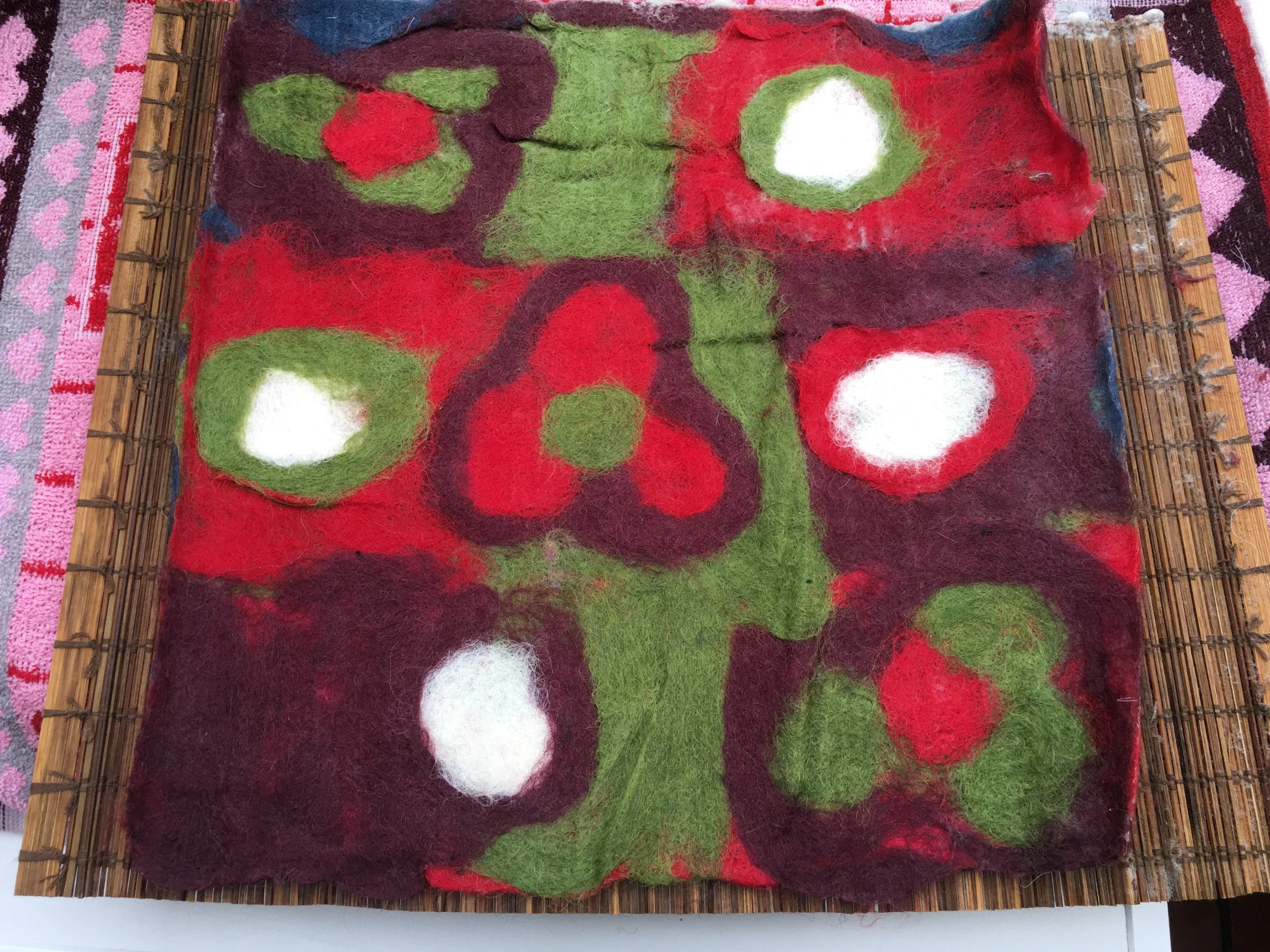 Colorful felted wool artwork with red, white, green, purple, and blue shapes, framed with bamboo sticks, and placed on a pink patterned rug.