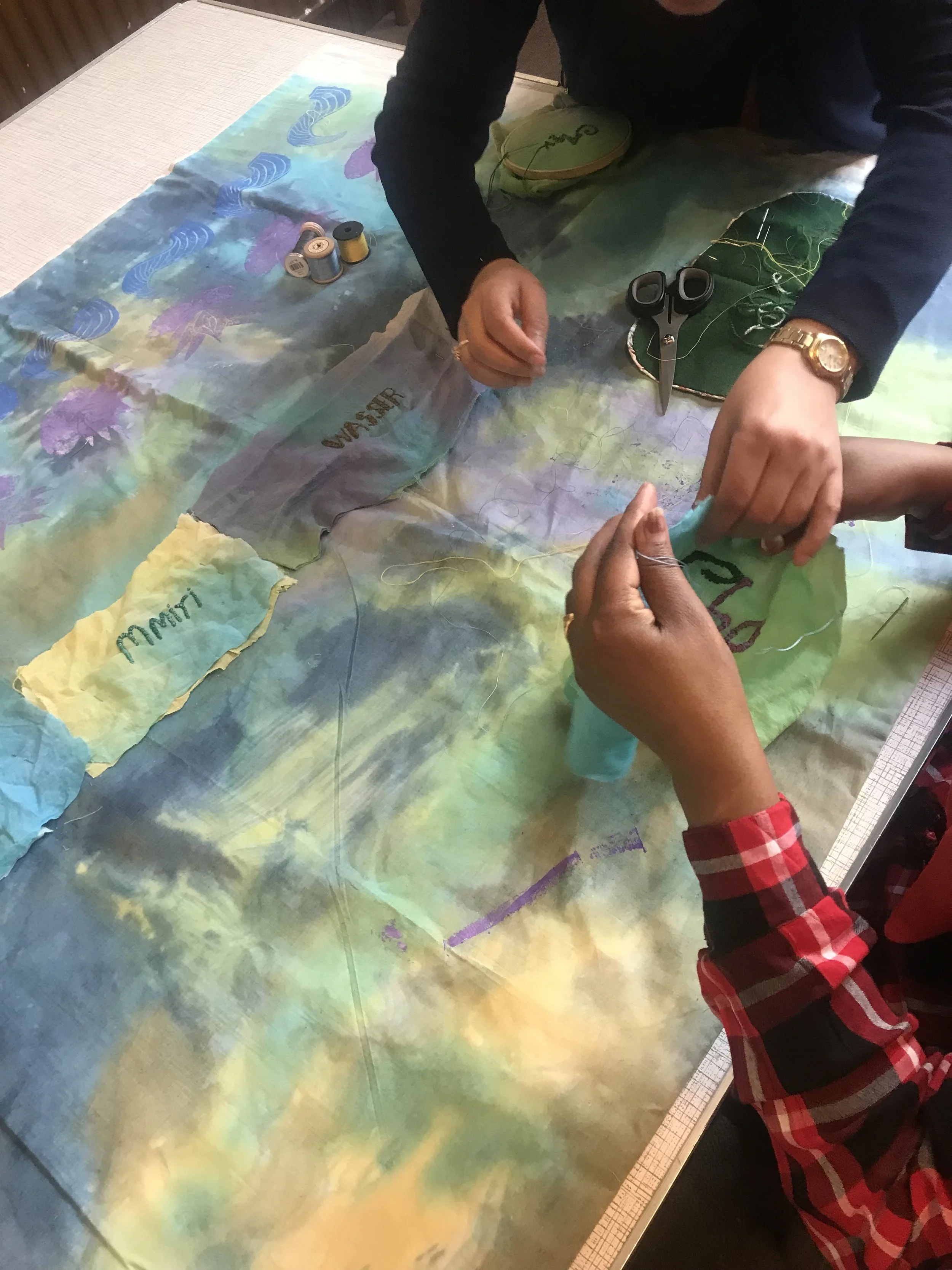 Close-up of two people sewing or embroidery on a colorful, artistic fabric with thread, scissors, and sewing supplies on the table.