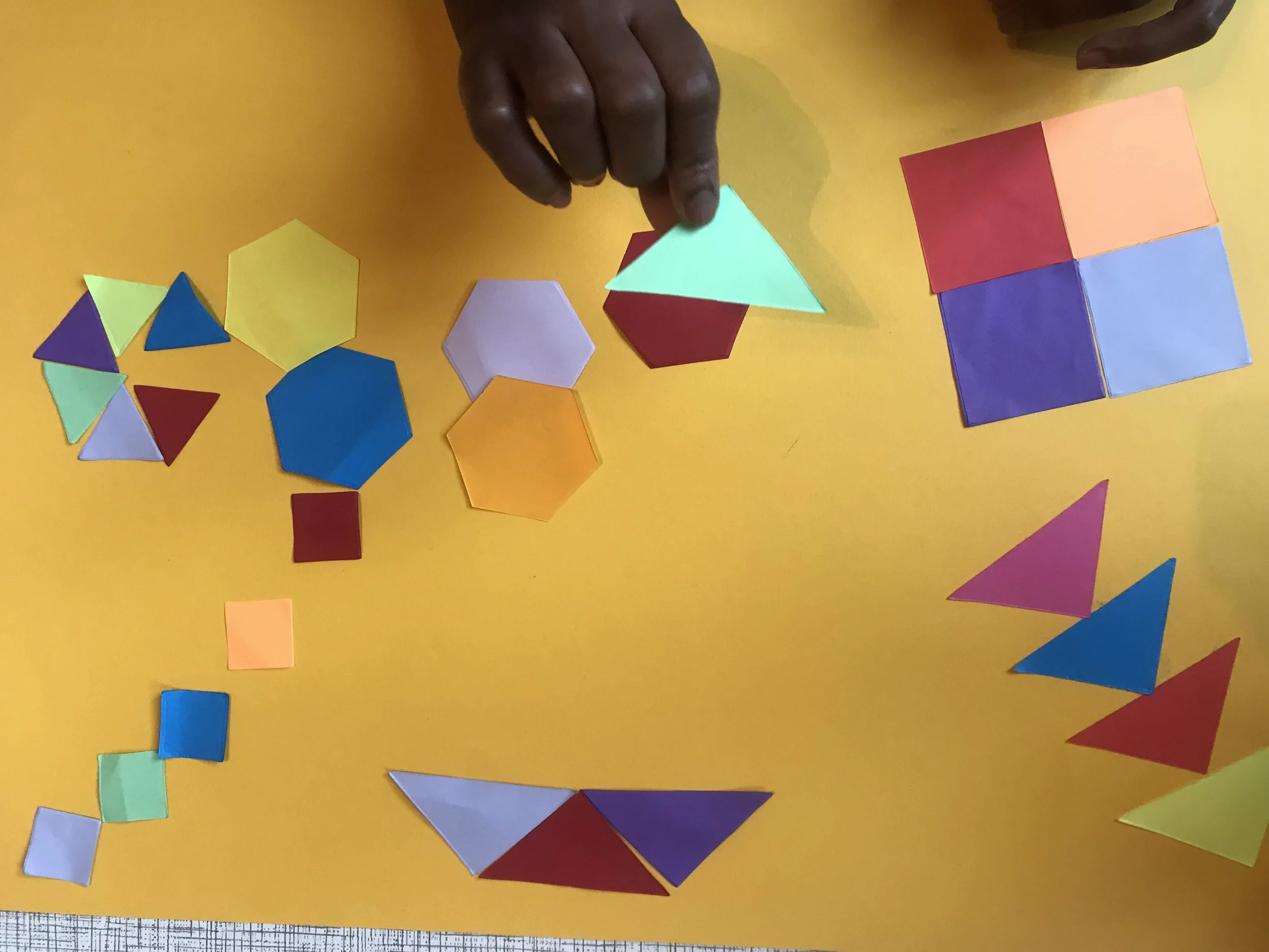 A hand with dark skin is arranging colorful geometric paper shapes on a yellow surface. The shapes include squares, triangles, and hexagons in various colors like red, blue, purple, orange, yellow, green, and white, some in stacks and some scattered.
