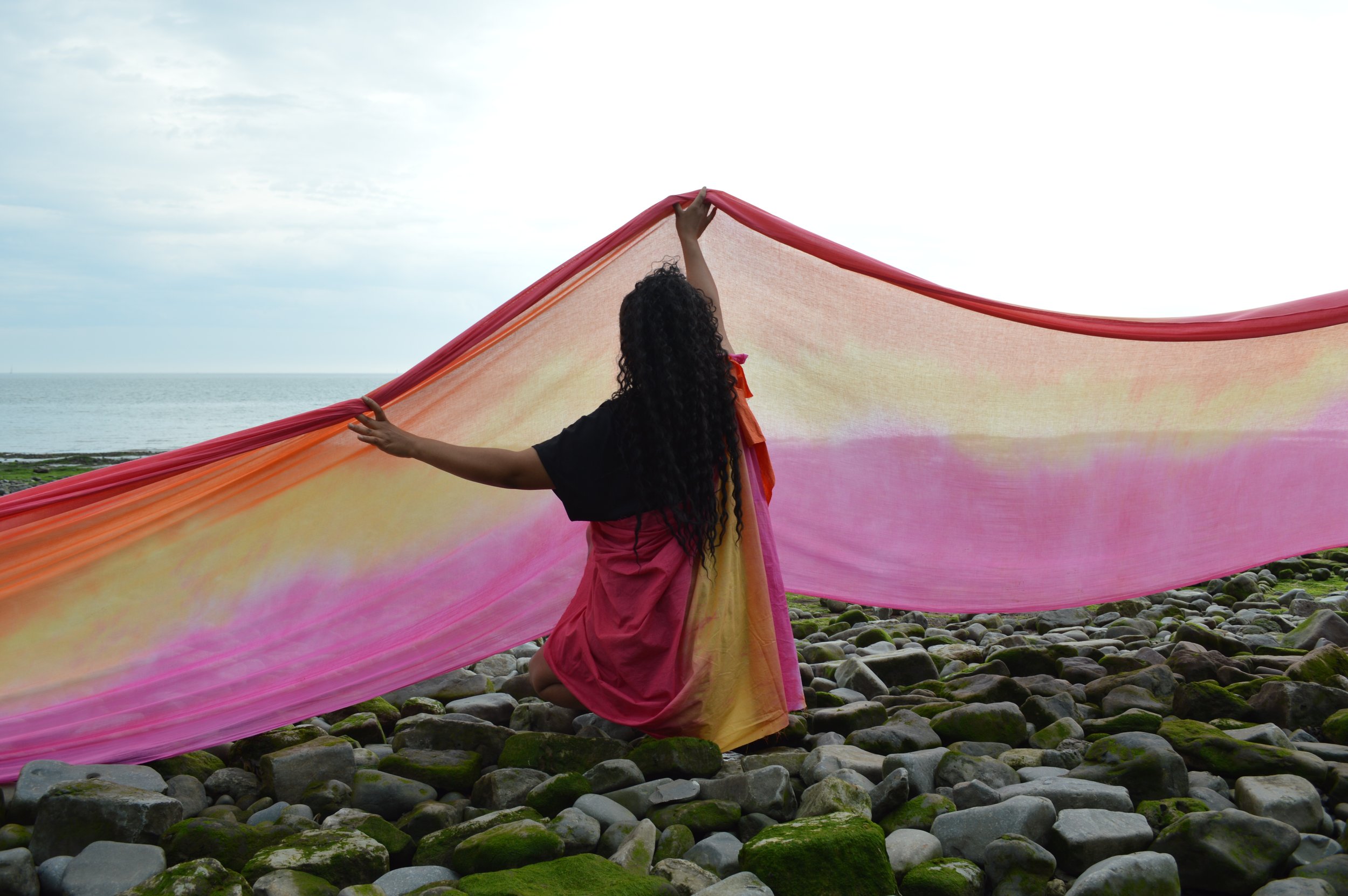 A woman in a pink sari holding a large colorful fabric near the rocky shoreline with the ocean and cloudy sky in the background.
