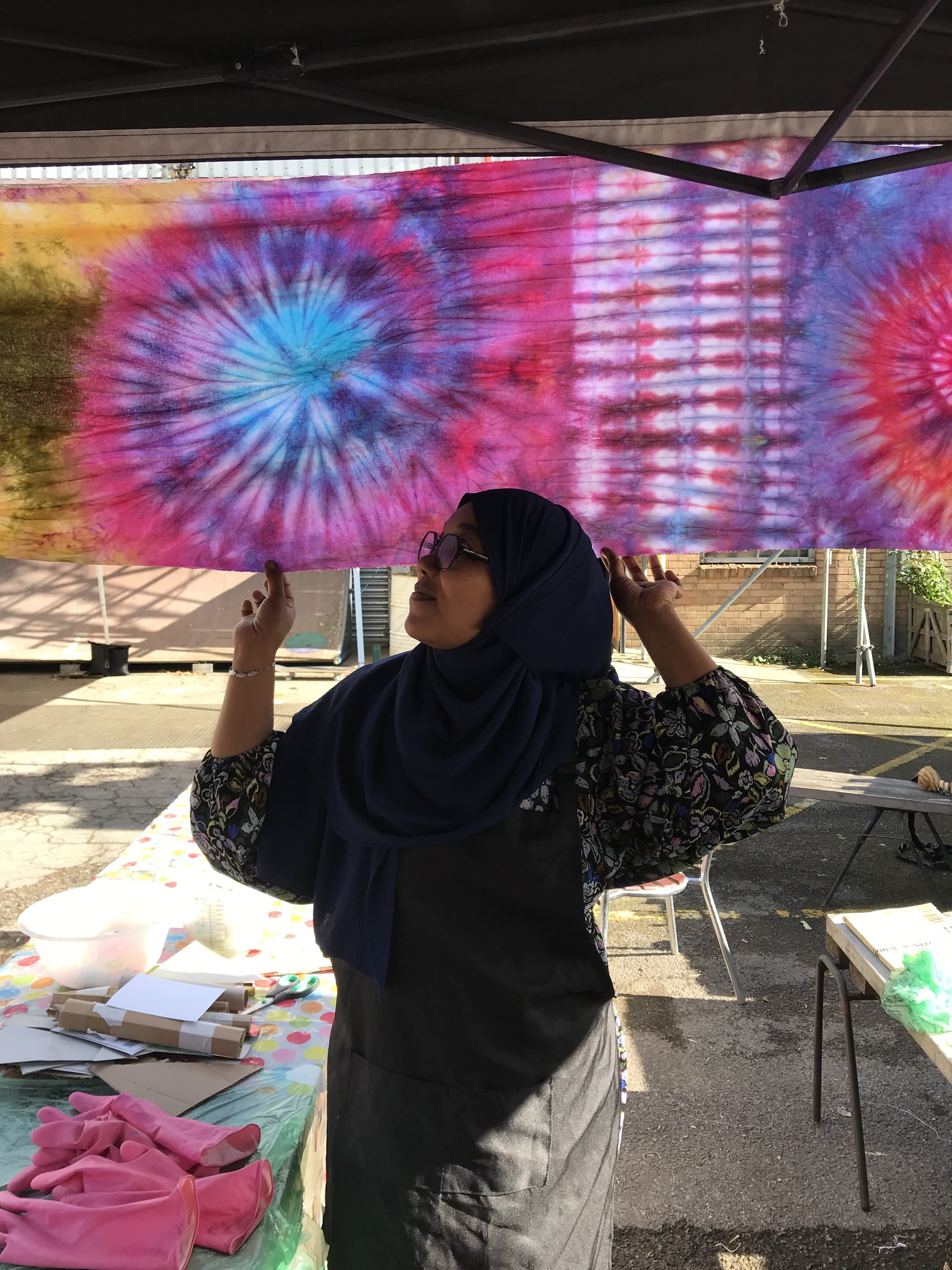 A woman in a dark hijab and glasses holding a tie-dyed fabric with vibrant pink, blue, and purple colors under a black canopy. She is working at a table covered with a colorful tablecloth, with various crafting supplies, outdoors with a brick building and trees in the background.