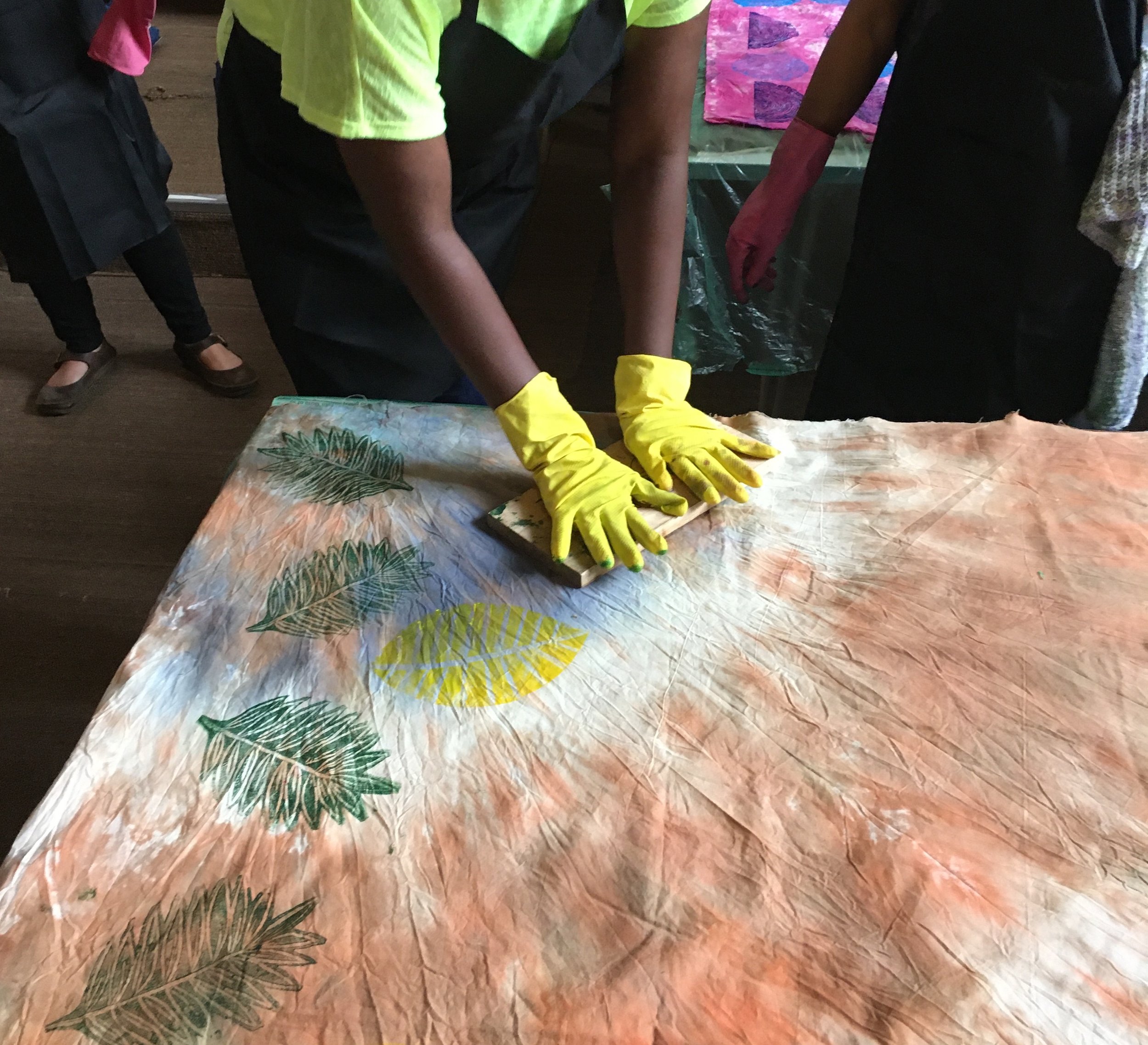 People wearing yellow gloves using a knife to cut a vegetable on a table covered with a leafy patterned tablecloth.