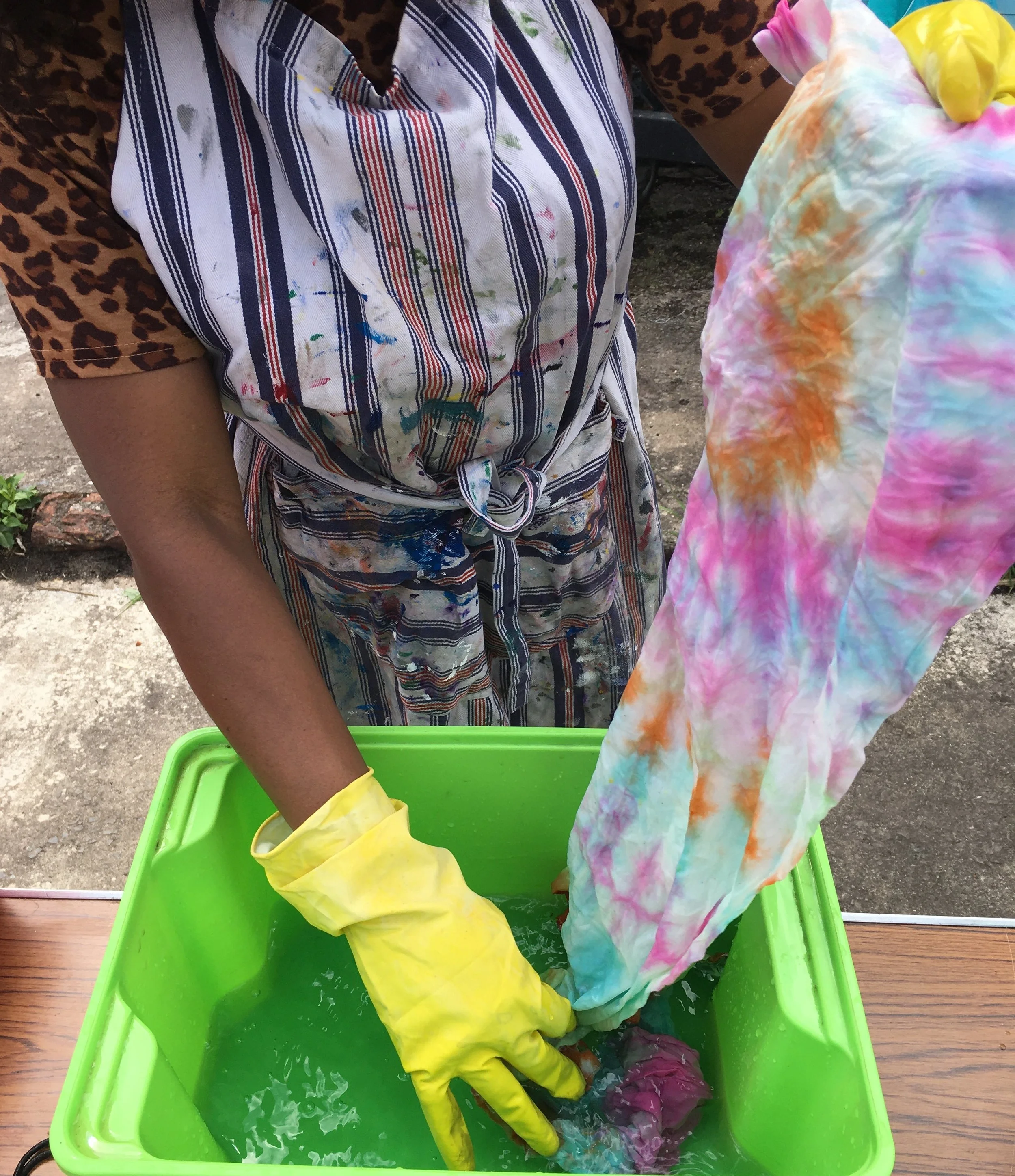 Child wearing yellow gloves holding colorful tie-dye fabric over a green container filled with water.