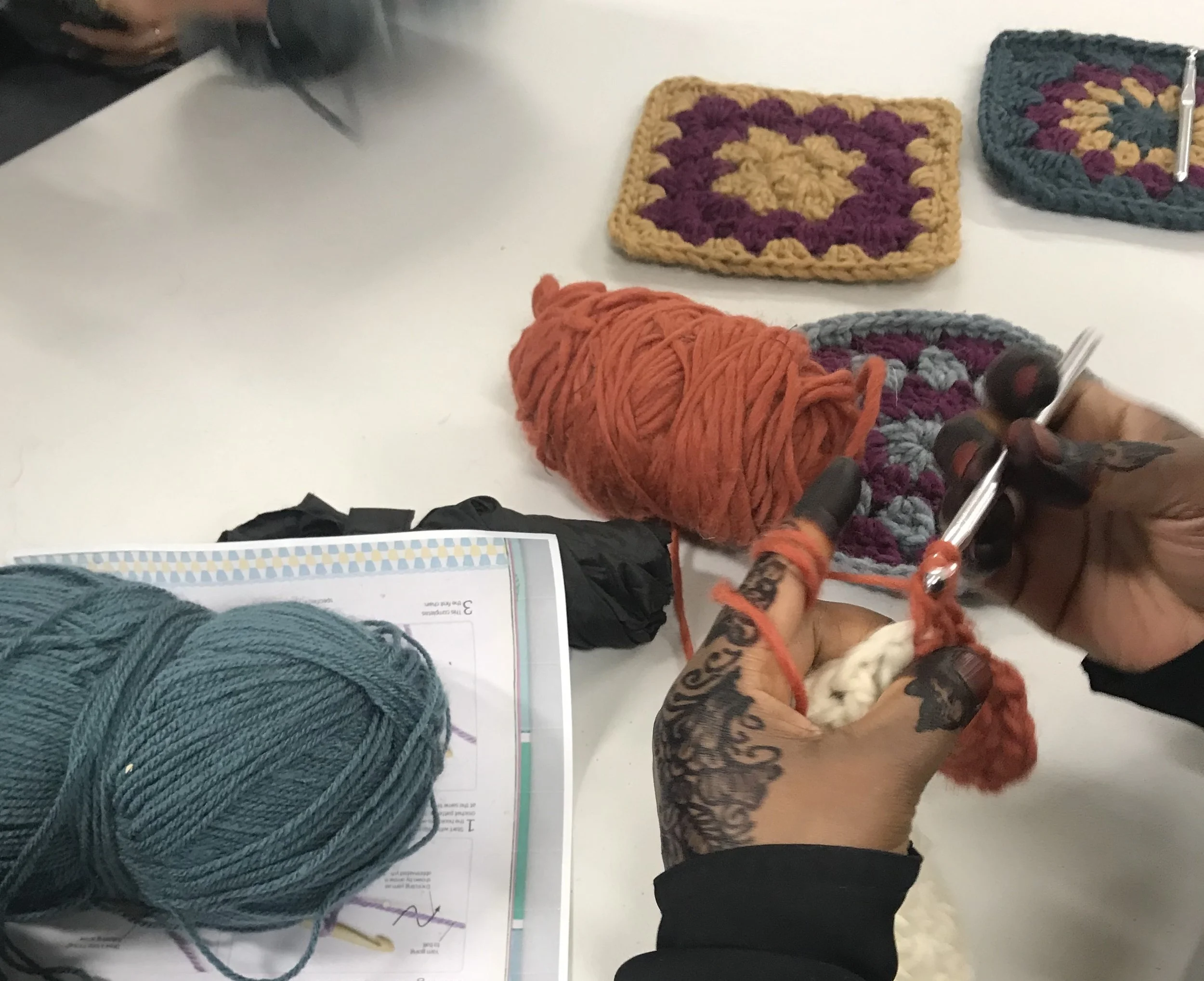 Close-up of a person's hands knitting with orange yarn, with skeins of teal and green yarn and crocheted pieces on a white table.