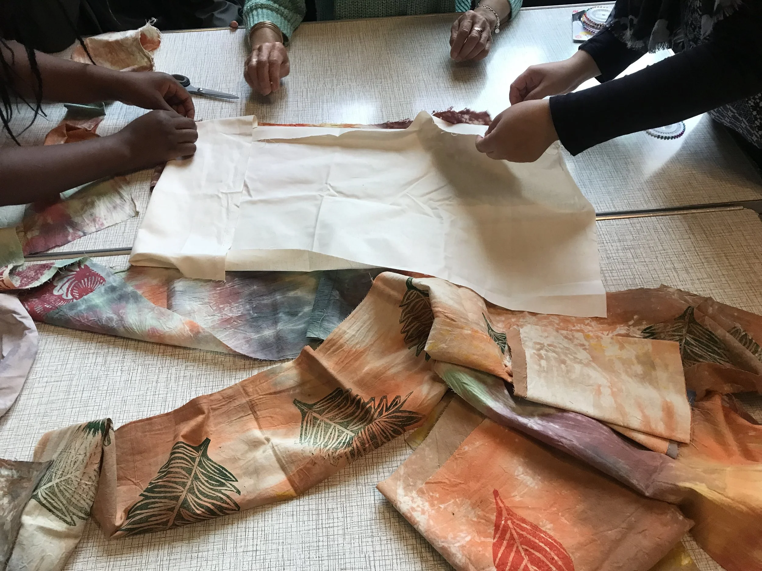 Hands of multiple people wrapping fabric or paper around a rolled item on a wooden table, with scissors and colorful fabric pieces nearby.