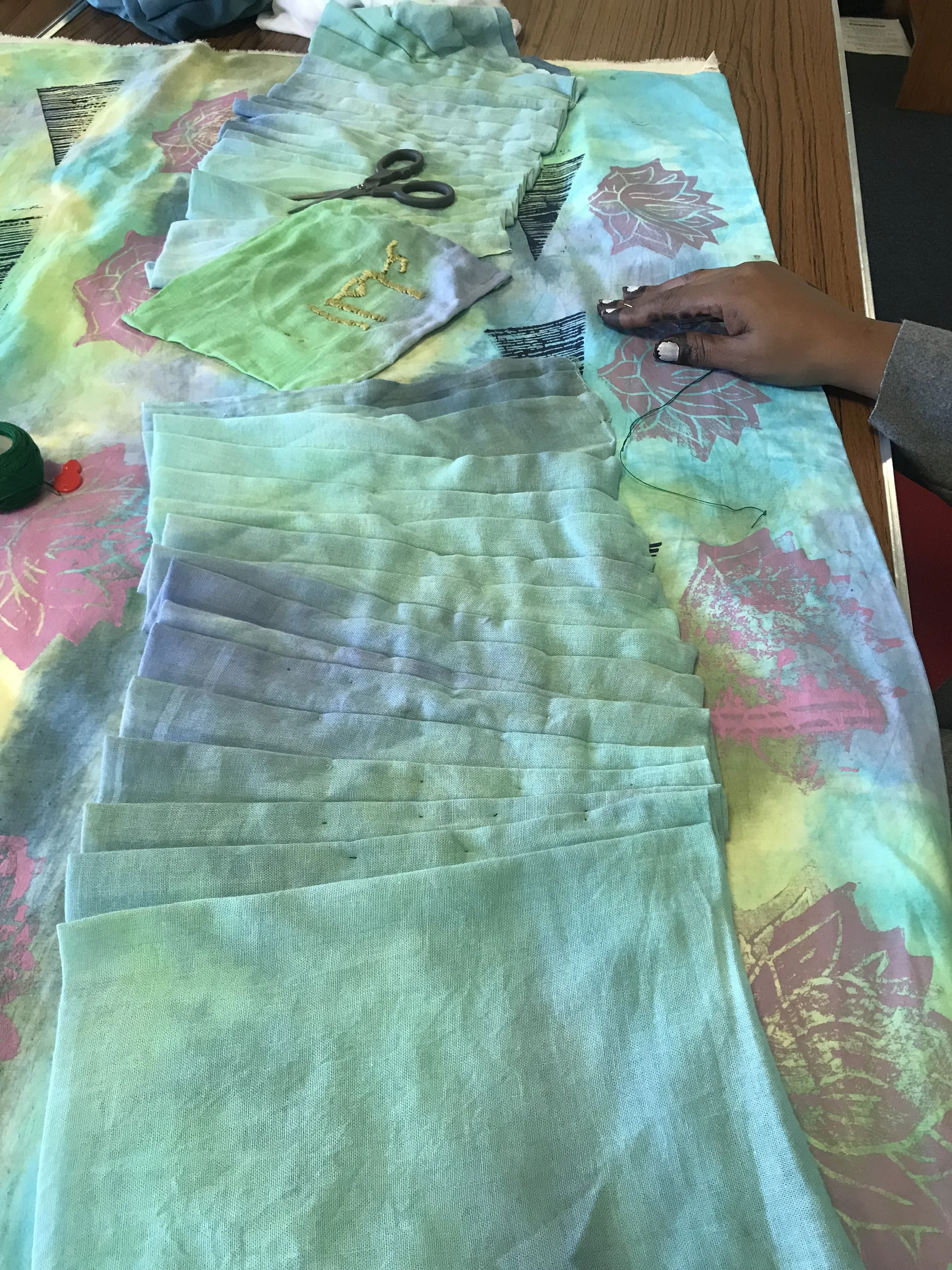 A person is sewing green fabric pieces on a table with a colorful tablecloth. The fabric pieces are stacked, and there are scissors and thread visible.