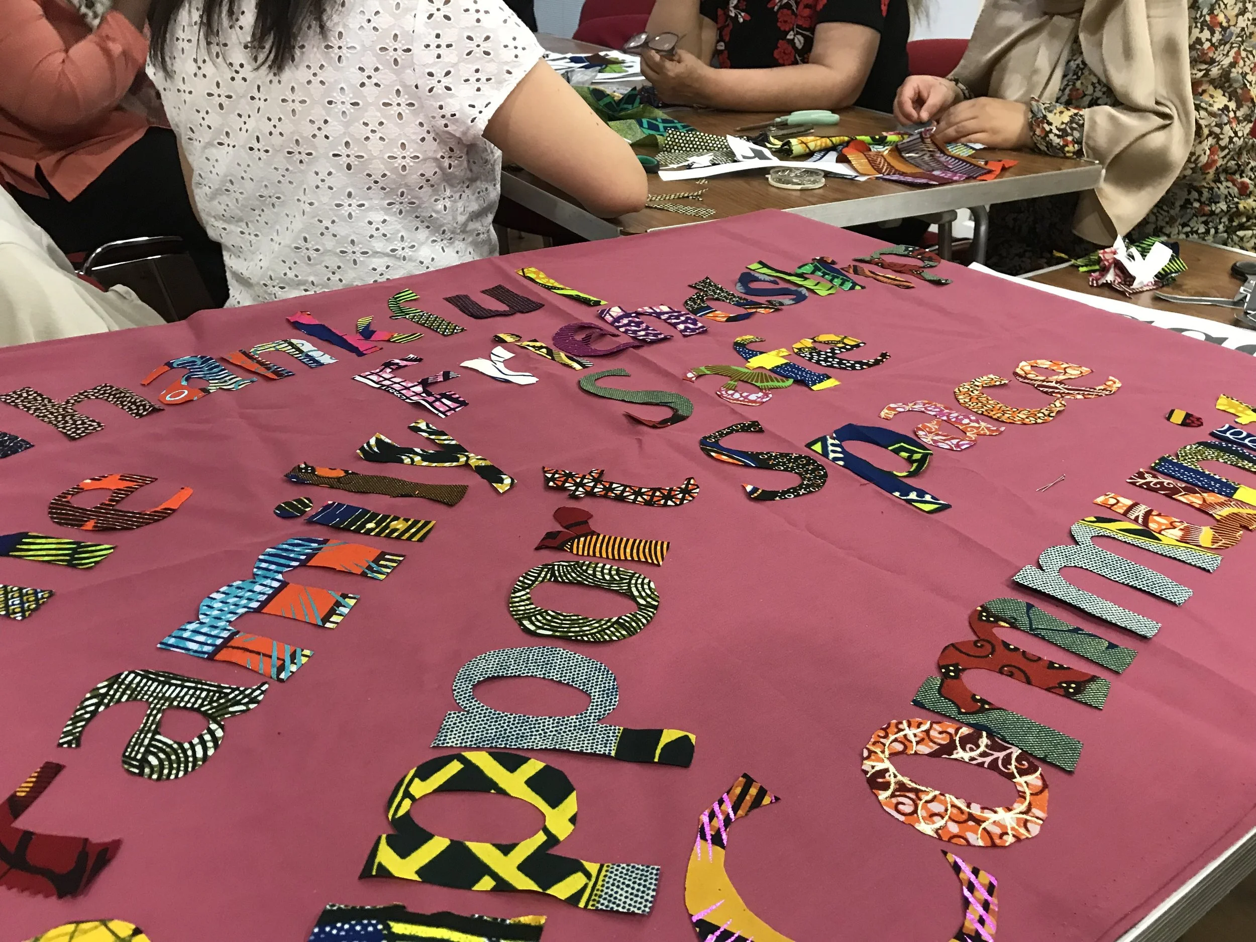 People gathered around a table working on a fabric craft project, with colorful fabric cutouts spelling out a message on a pink tablecloth.