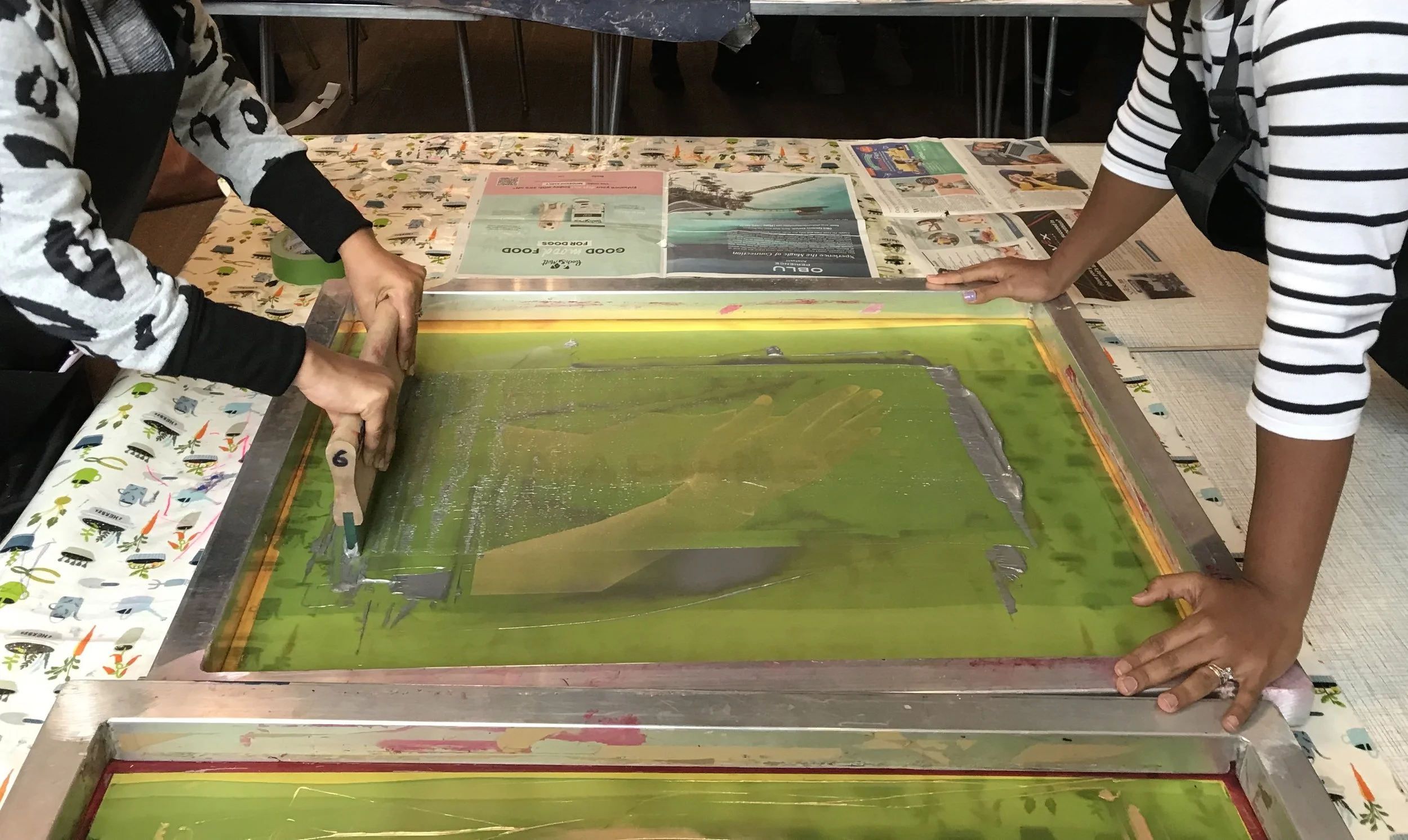 Two people are pressing handprints onto a green ink pad on a table covered with a colorful tablecloth, possibly for a handprint art project or event.