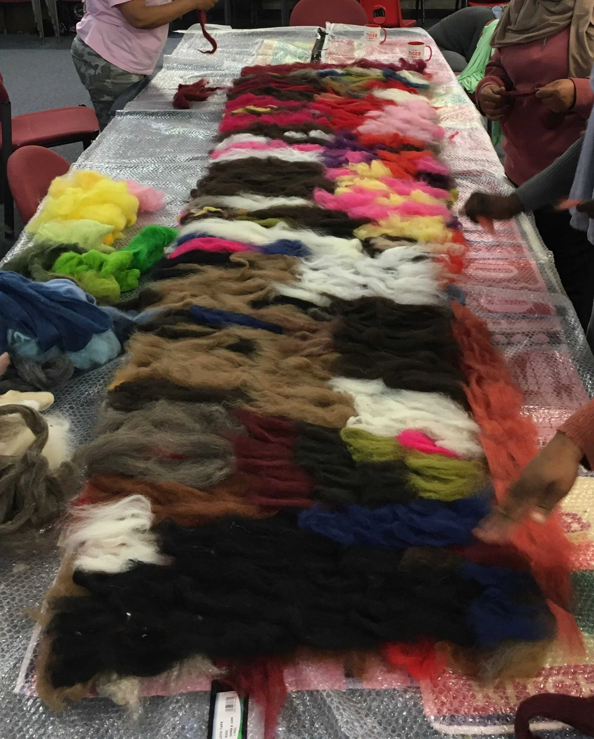 Table covered with colorful wool fibers, surrounded by people, some holding or tending to the fibers, with red chairs and cups in the background.