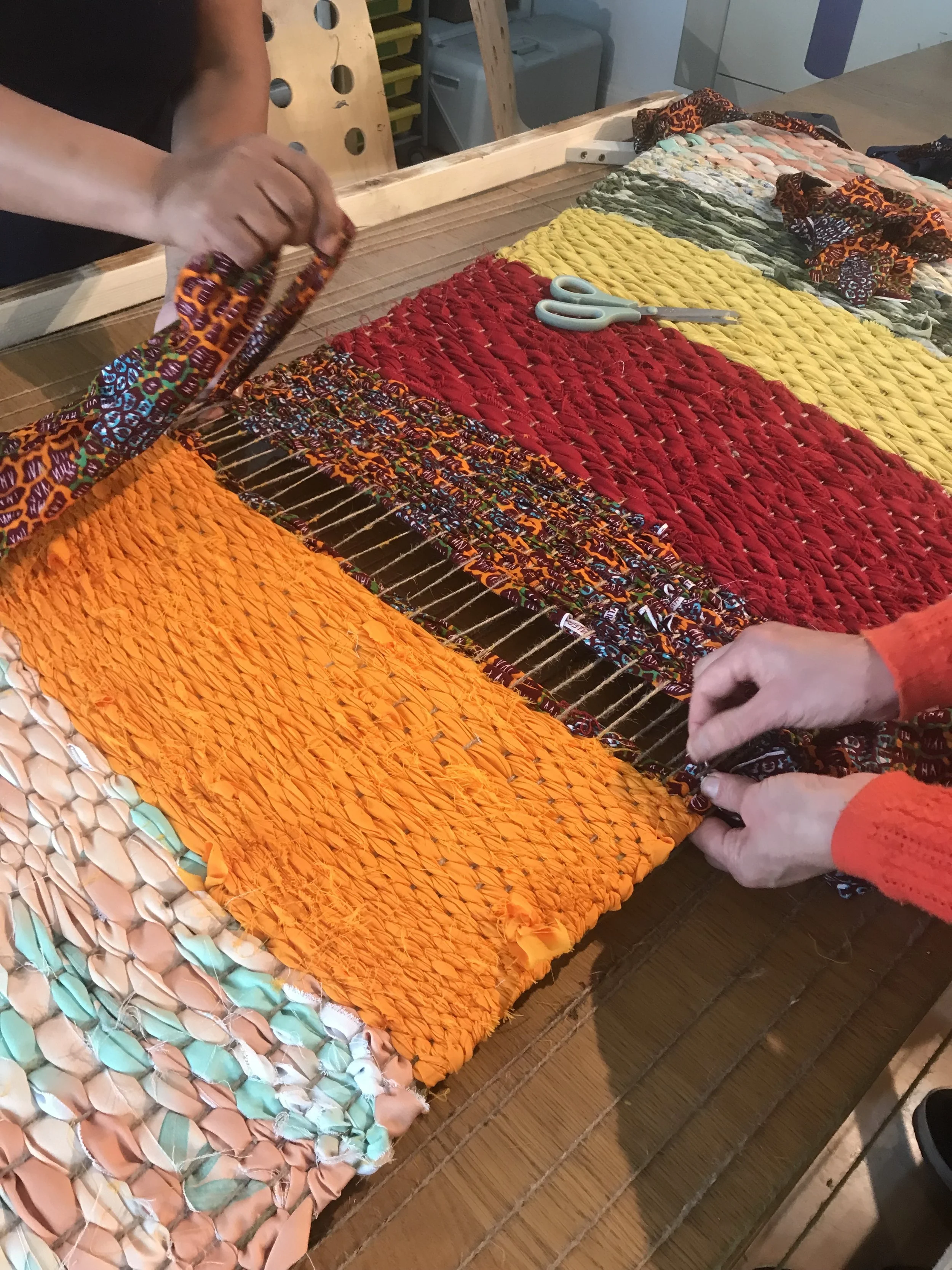 People weaving a colorful rag rug with fabric strips on a loom.