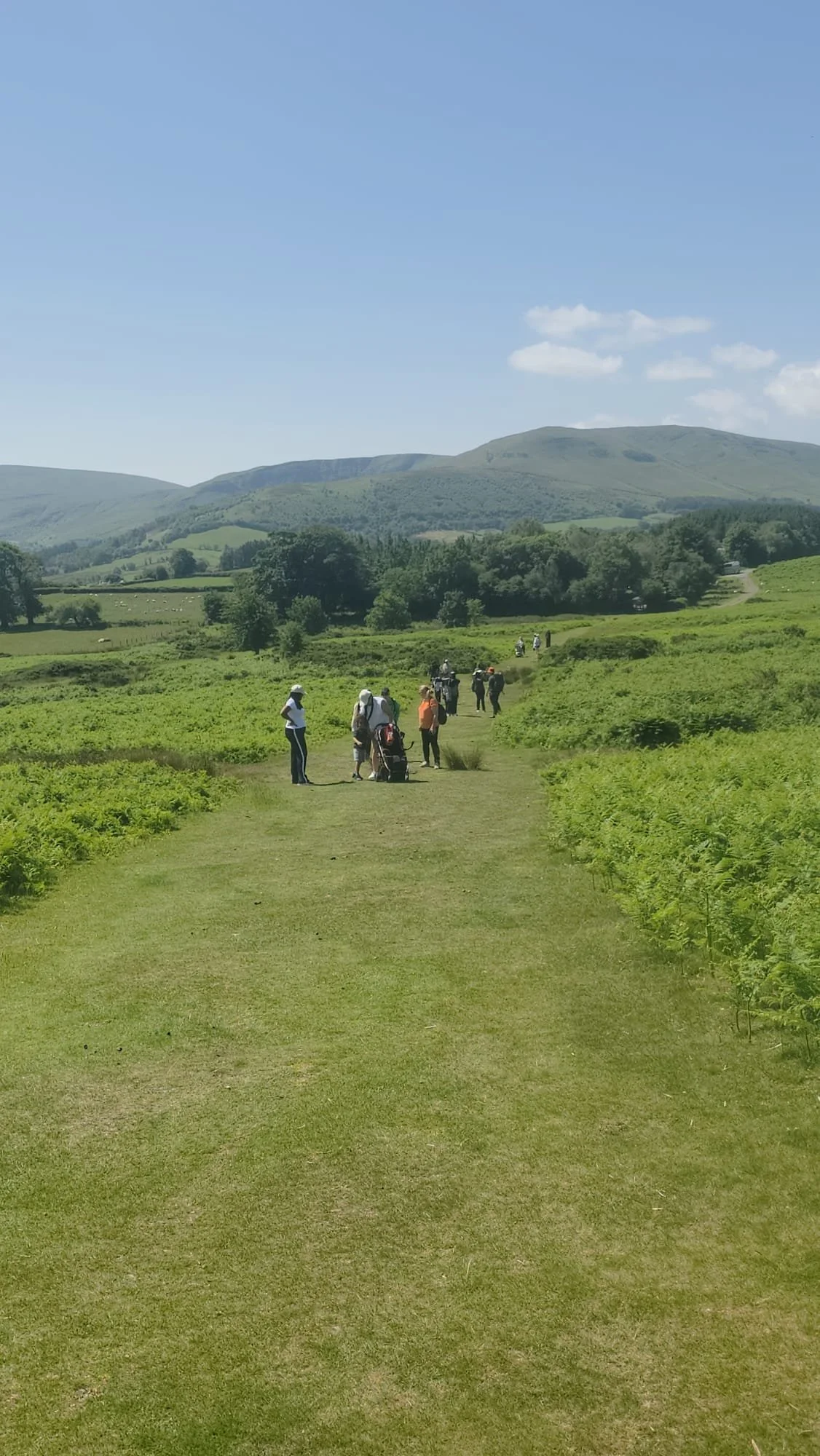 People walking along a grassy path through green fields with hills in the background under a blue sky with a few scattered clouds.