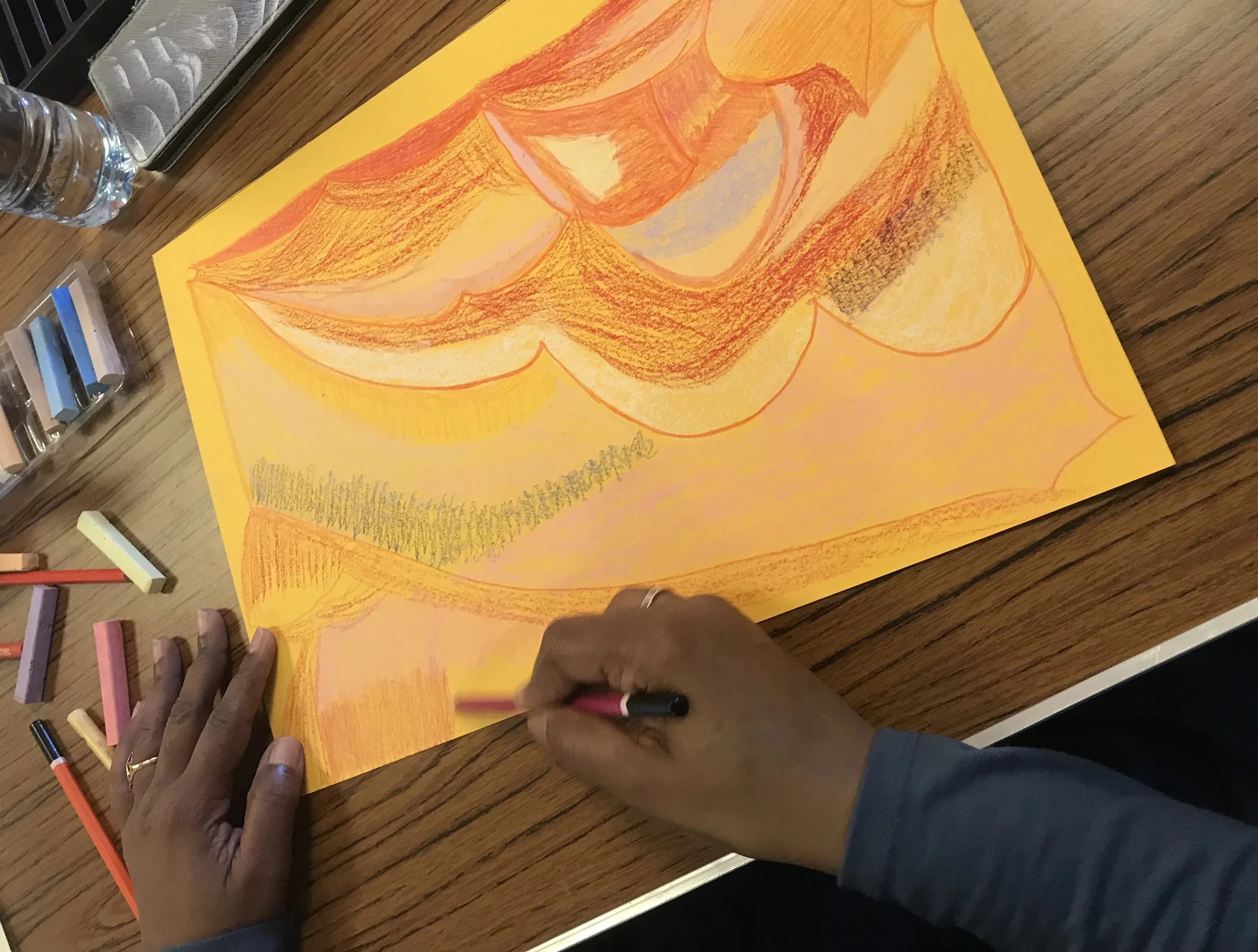 A colorful drawing of a pumpkin on yellow paper, with people using colored pencils and markers on a wooden table.