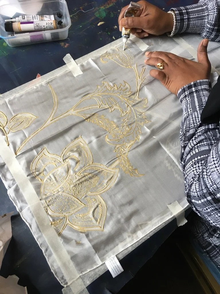 Person creating with gold leaf, floral design on fabric, supplies in container nearby.