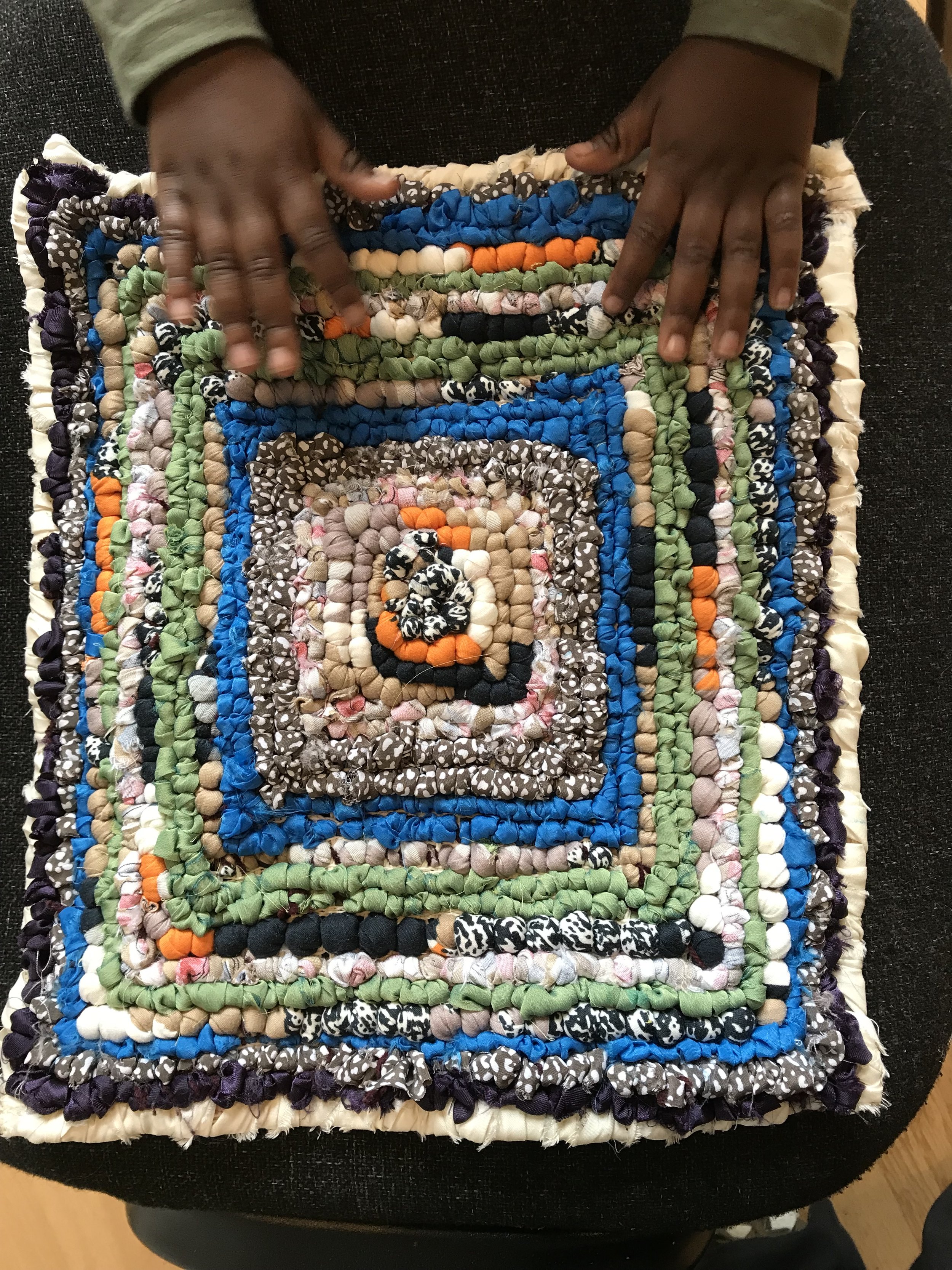 Colorful braided fabric placemat in various colors including green, blue, orange, beige, black, and white, with a black and white polka dot pattern, placed on a dark surface.