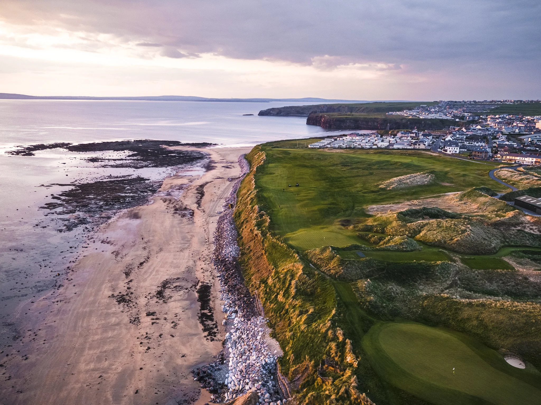 An aerial view of a coastal town with a beach, cliffs, and a golf course at sunset.