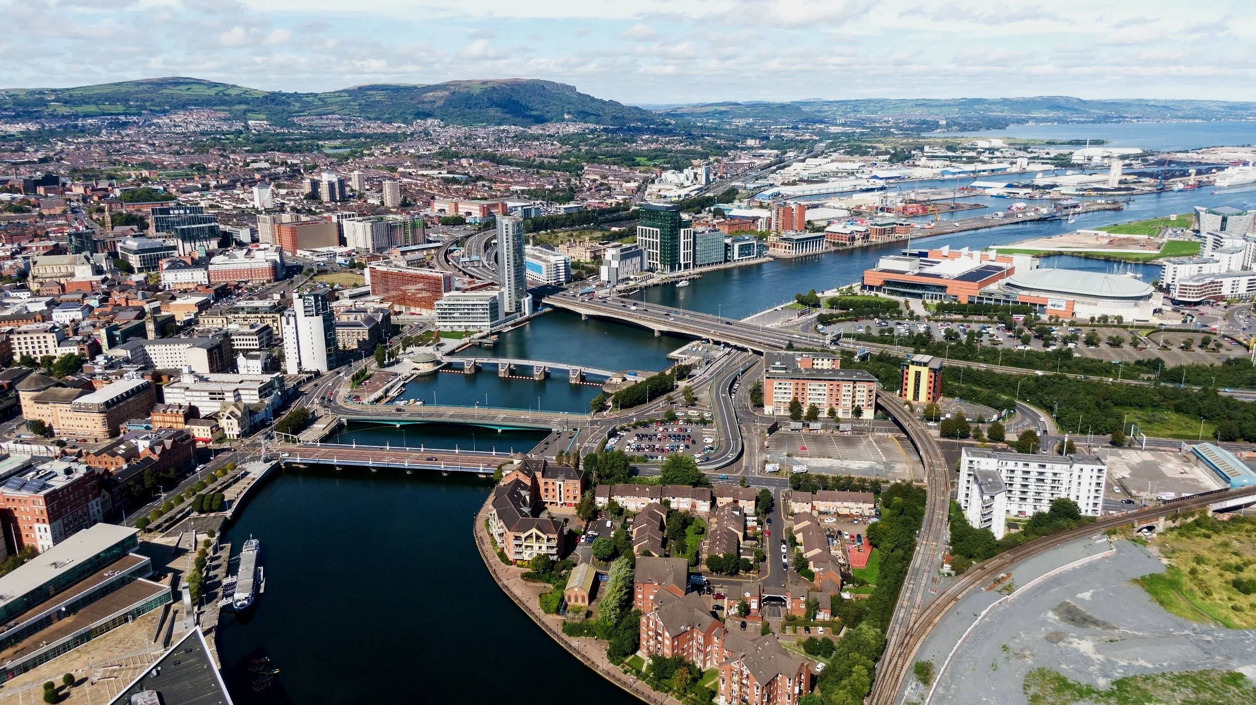 Aerial view of a city with a river running through it, surrounded by bridges, buildings, and green hills in the background.