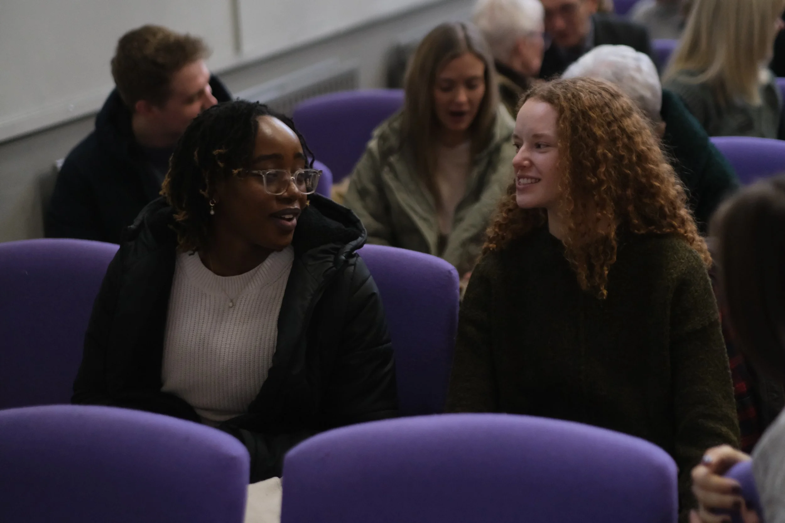 Group of diverse people sitting in an auditorium, engaging in conversation and smiling.