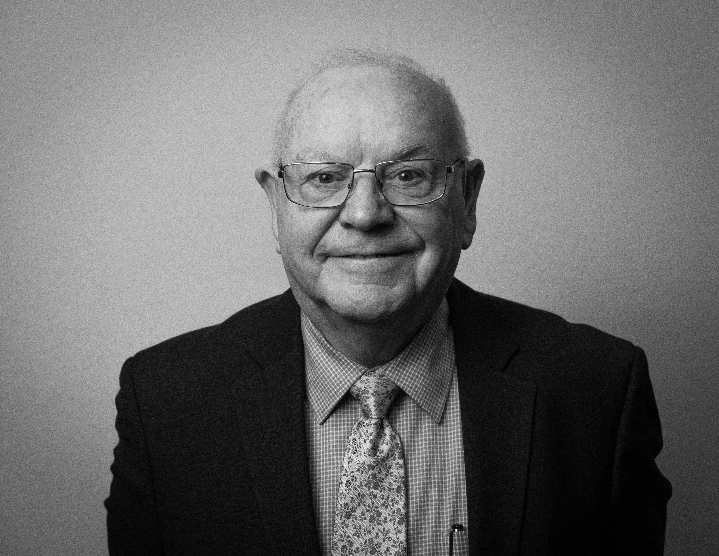 Black and white photo of an elderly man with glasses, smiling, wearing a suit, a patterned tie, and a collared shirt.