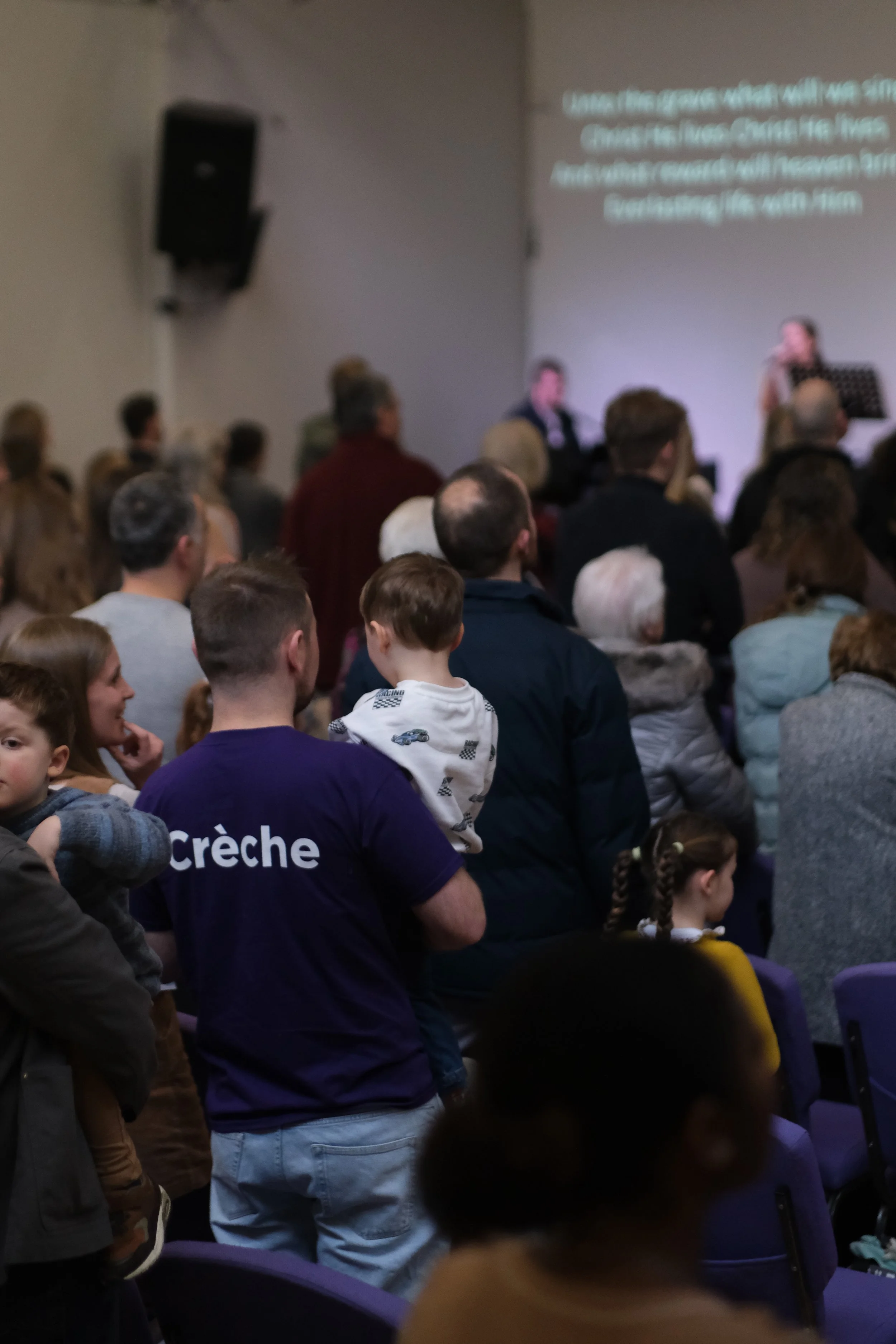 A gathering of children and adults seated and standing in a room, watching a presentation projected on the wall. The text on the screen is partially visible. The room appears to be a classroom or conference space.