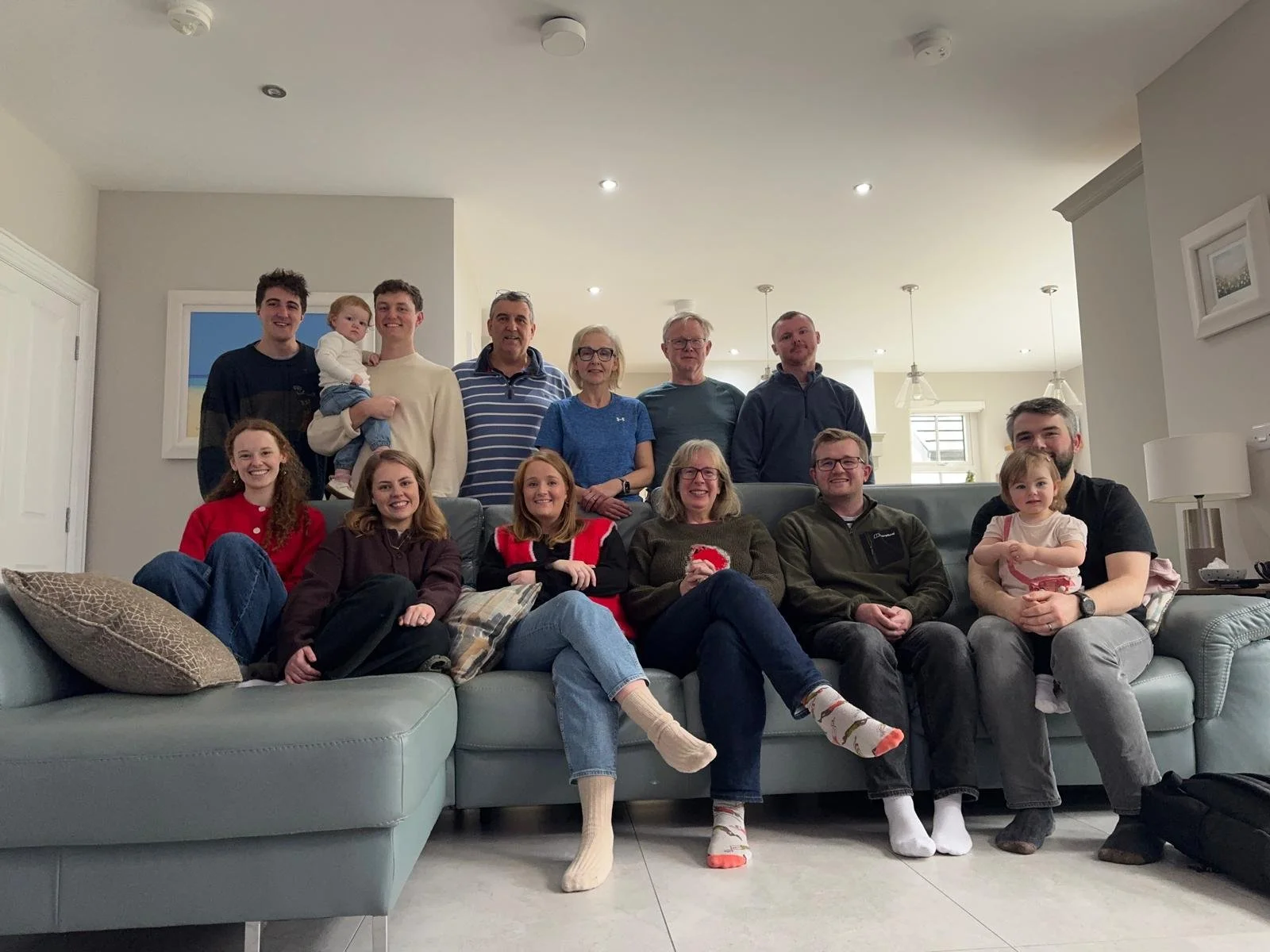 A multi-generational family group portrait in a living room, with the family members sitting on a sofa and standing in the background, smiling for the photo.