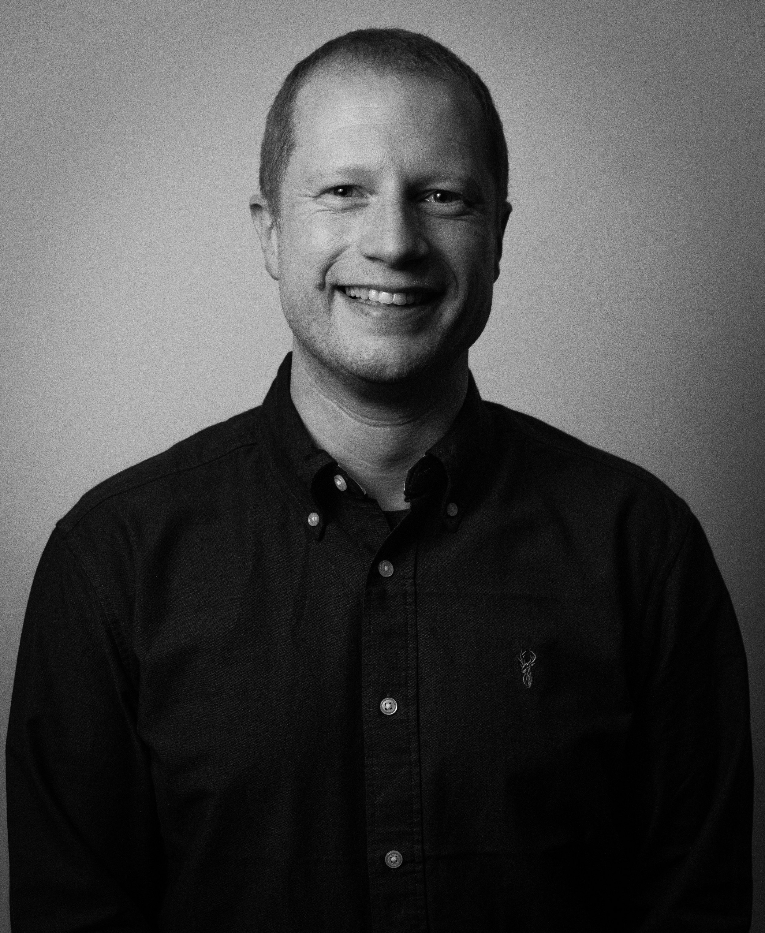 A black and white portrait of a smiling man wearing a dark button-up shirt with a small embroidered logo of a deer on the chest.
