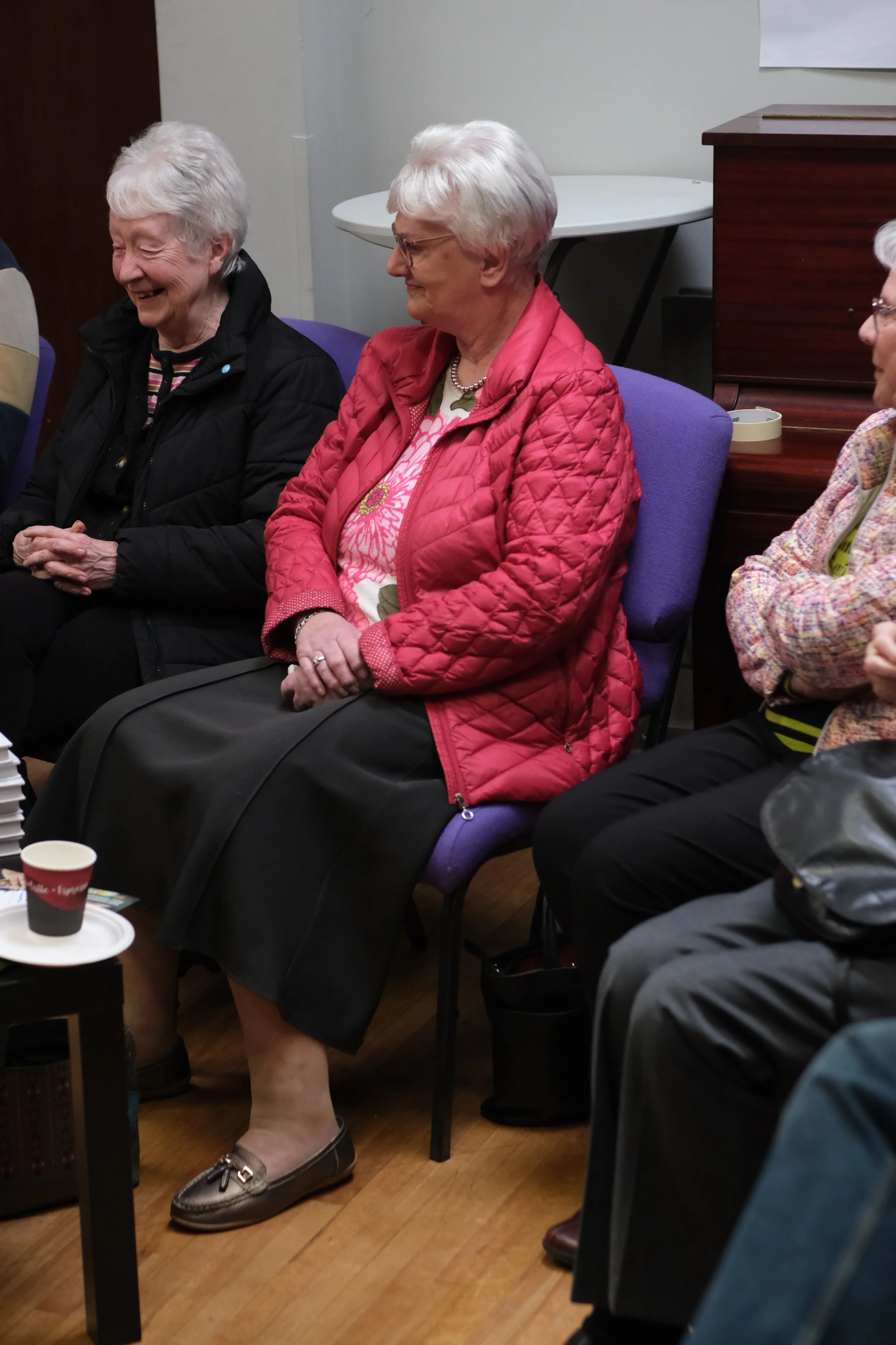 A group of elderly women sitting in chairs in a room. They appear to be engaged in conversation and are smiling. The woman in focus is wearing a red quilted jacket, a floral patterned top, a pearl necklace, and has short white hair. There are cups and papers on the table in front of them.