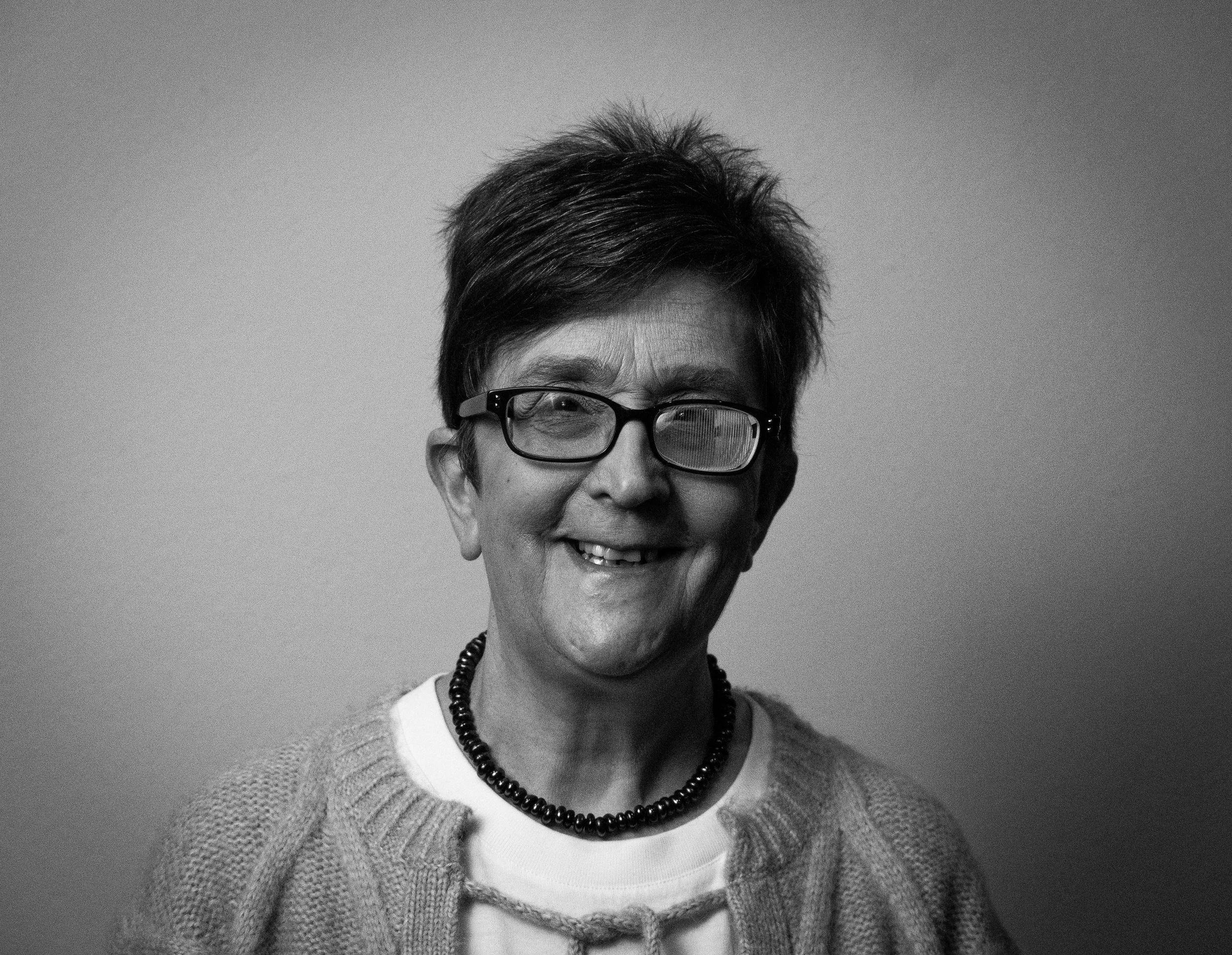 A black and white portrait of an elderly woman wearing glasses, a string of beads, a blazer, and a white shirt, smiling against a plain background.