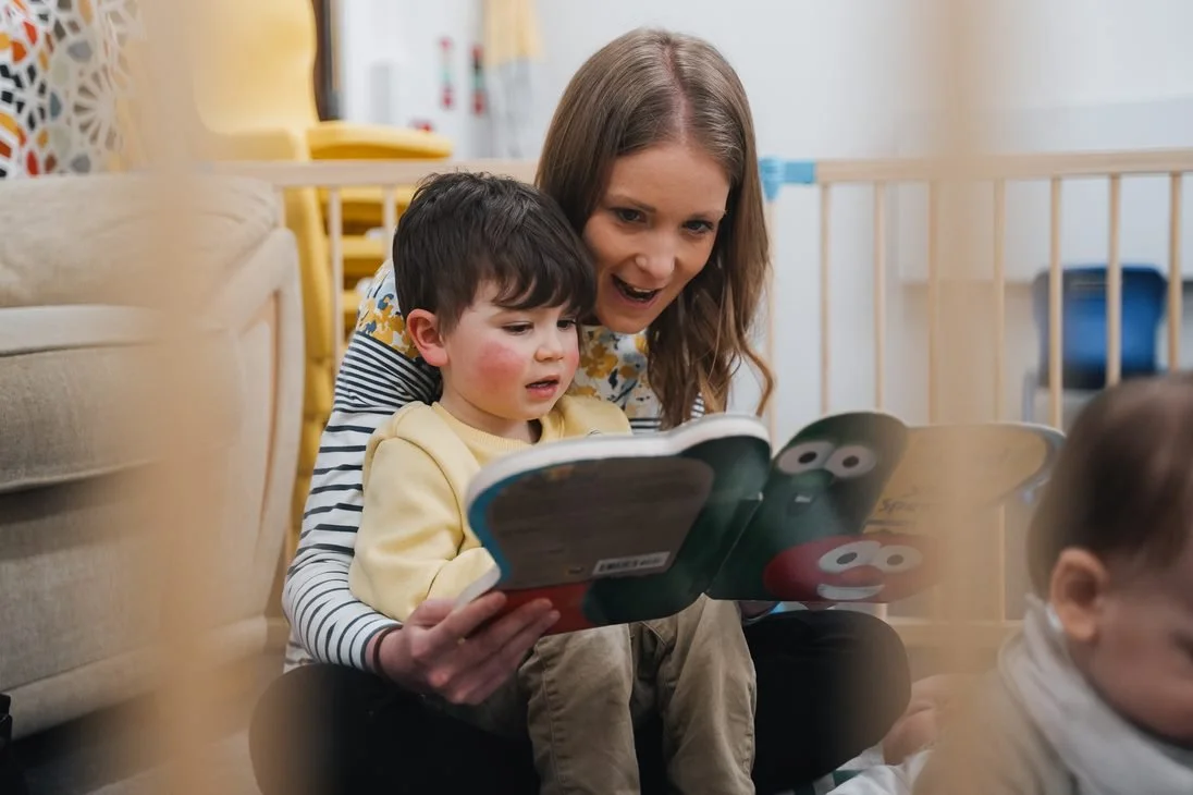A woman and a young boy sitting on the floor, looking at a colorful children's book together in a cozy room.