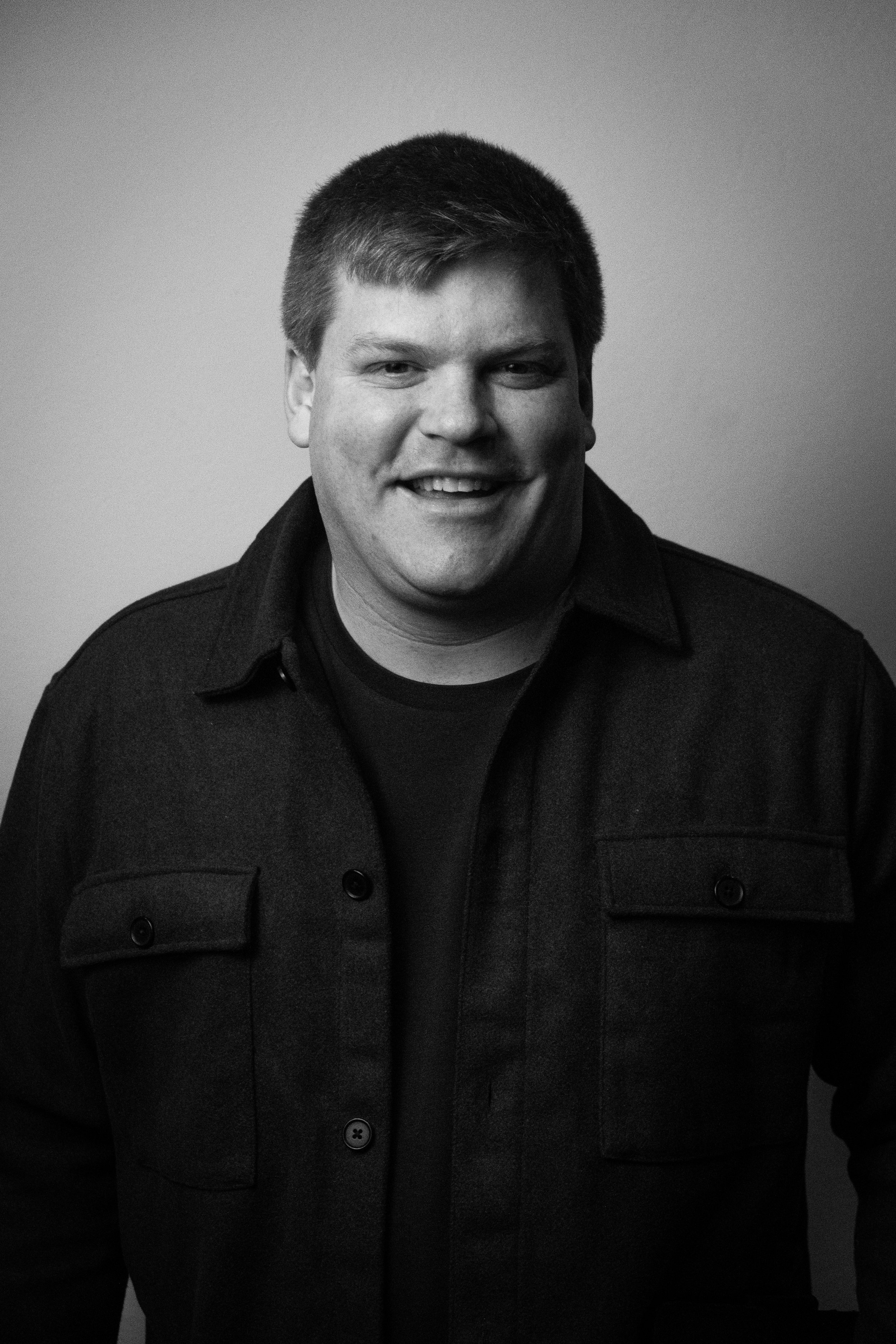 Black and white photo of a smiling man with short hair wearing a dark buttoned shirt and a dark coat, standing against a plain background.