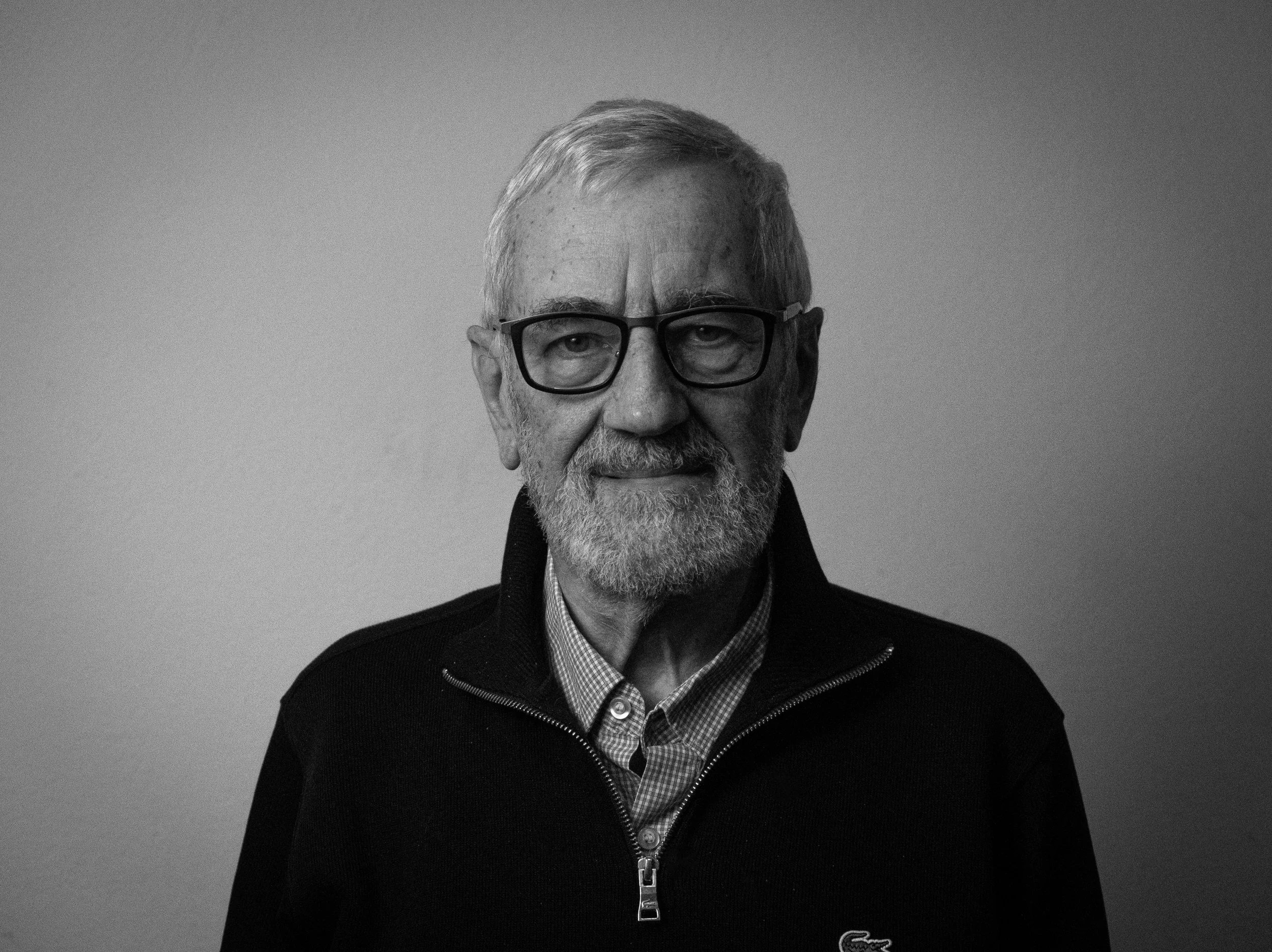 A black and white portrait of an elderly man with glasses, a beard, and gray hair, wearing a zipped black sweater over a collared shirt, against a plain background.
