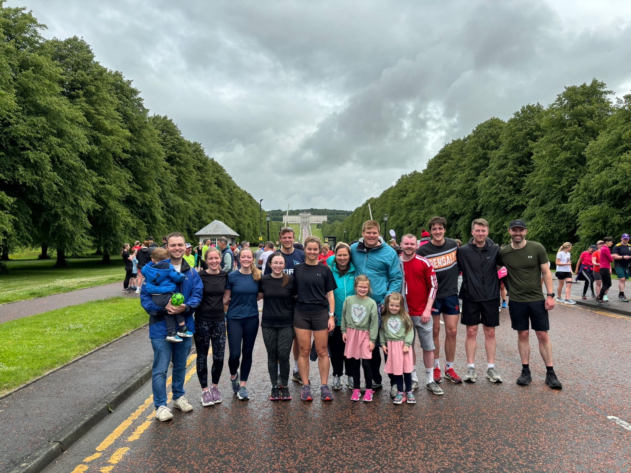 A group of smiling people standing on a wet street in front of a large park, with trees lining both sides and a historical building visible in the background under a cloudy sky.
