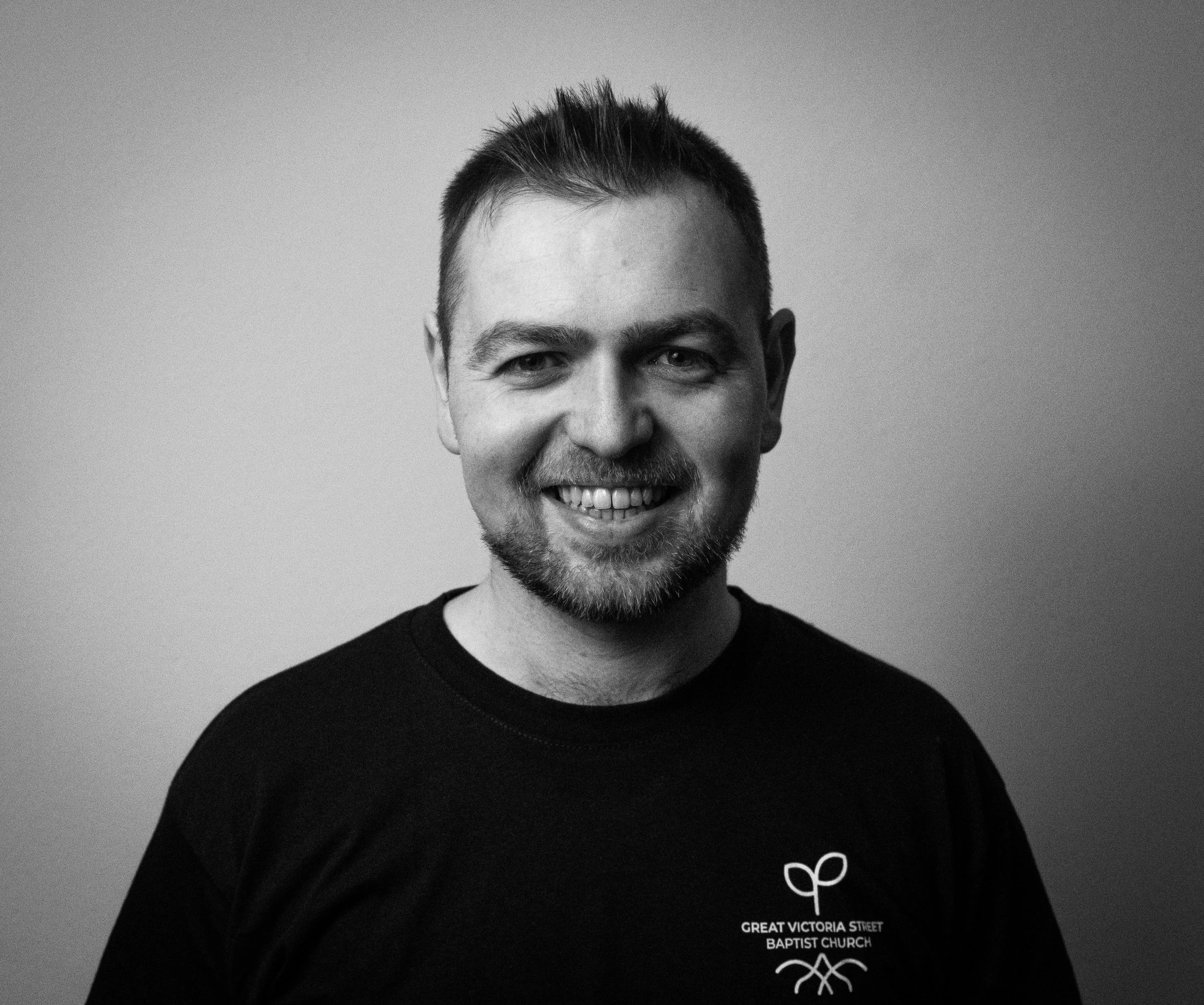Close-up of a smiling man with short spiked hair wearing a black T-shirt with white text and a logo, standing against a plain background.