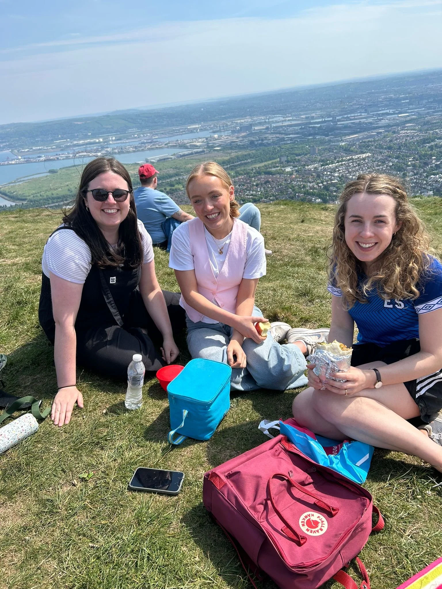 Three young women sitting on a grassy hilltop with a scenic view of a city and river below, enjoying a picnic on a sunny day.