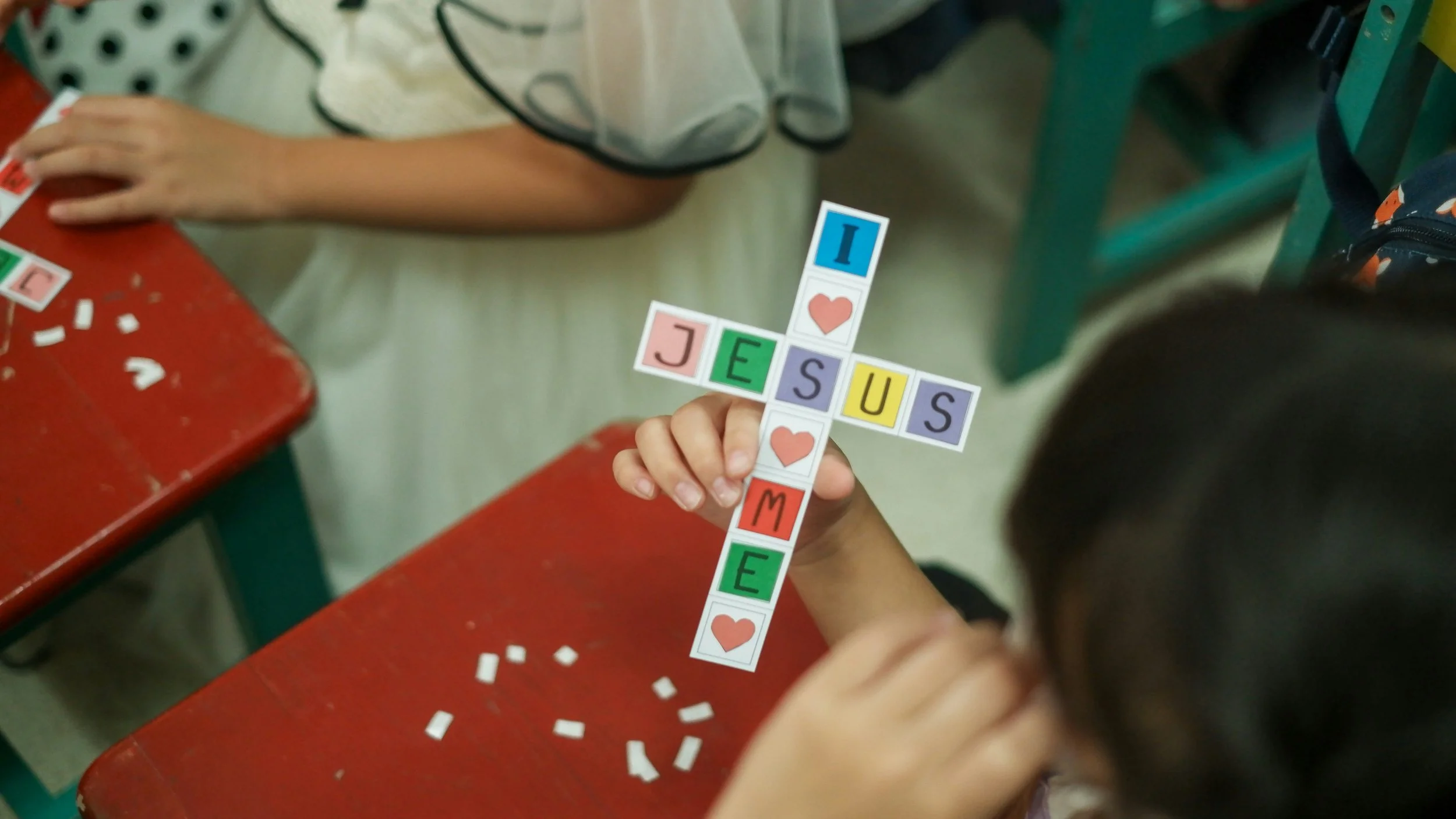 Child holding a crossword-style display spelling 'JESUS' with colorful letter tiles and hearts, in a classroom setting.