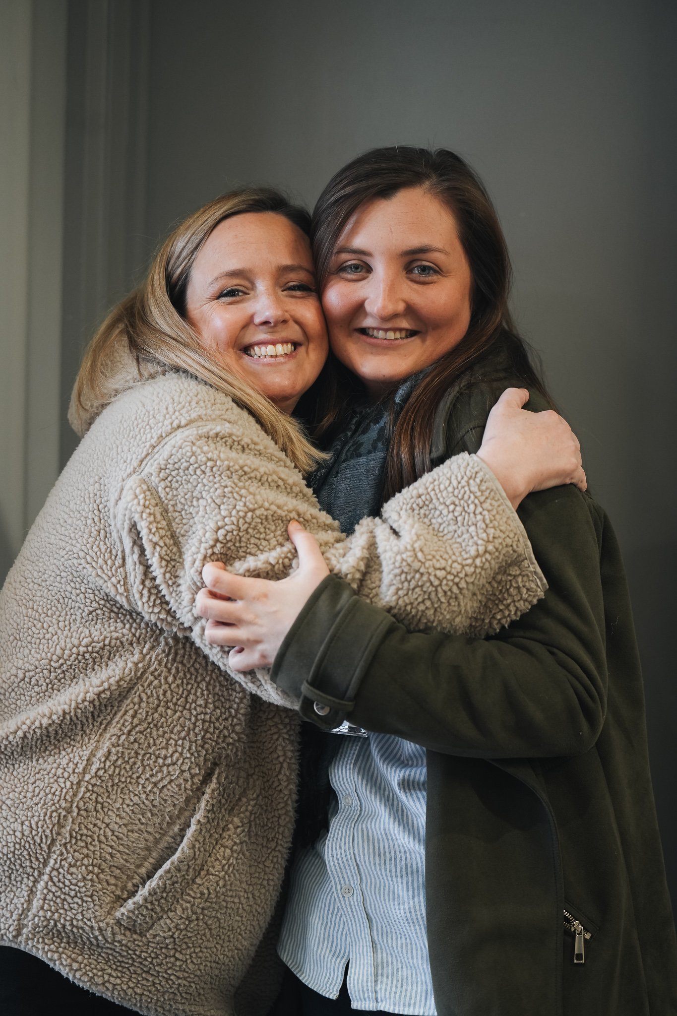 Two women hugging each other and smiling at the camera. One woman is wearing a beige fuzzy coat, and the other is wearing a dark jacket over a striped shirt. They are standing against a plain grey wall.