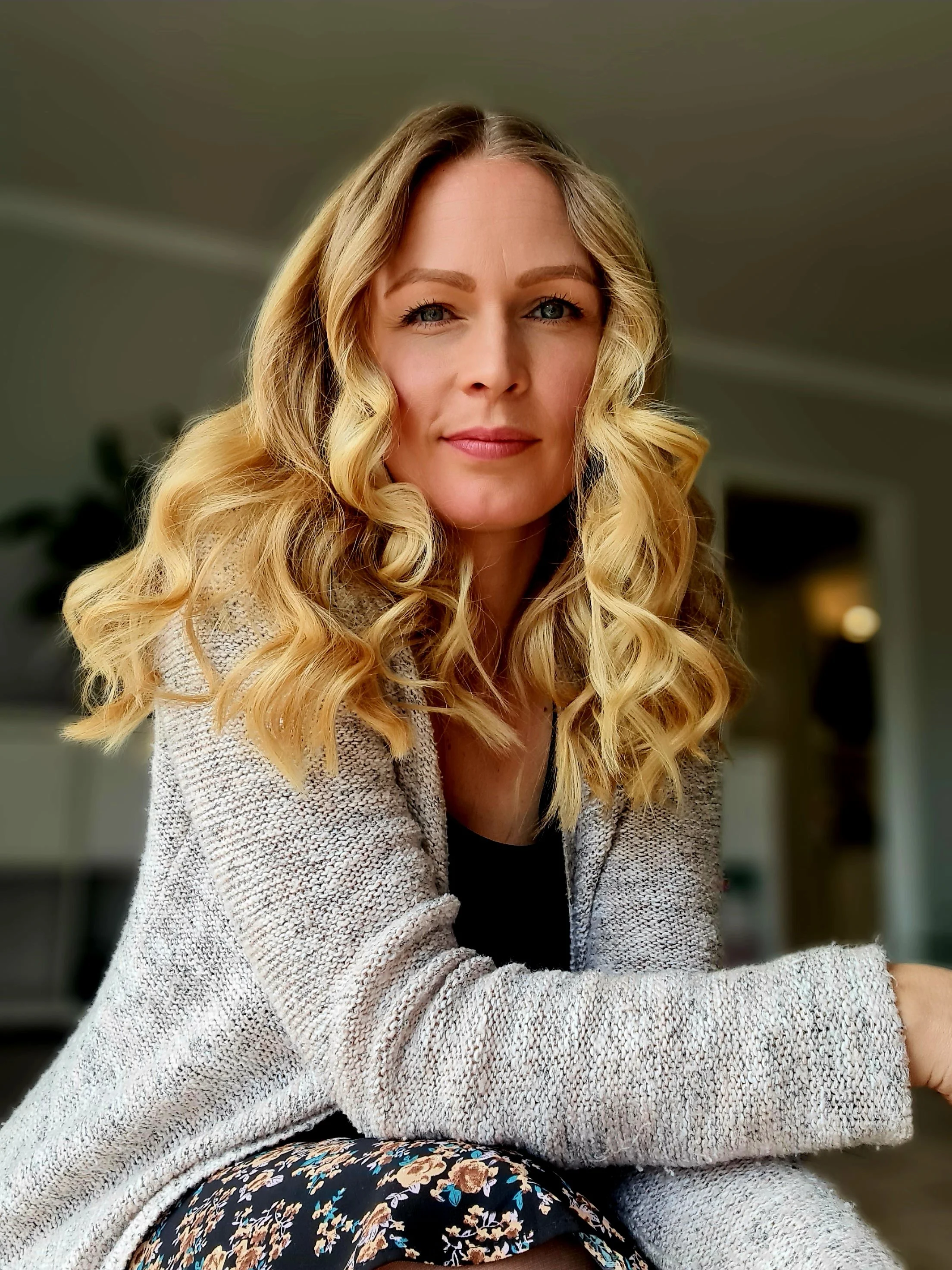 Eine Frau mit langen, lockigen blonden Haaren sitzt in einem Raum und blickt in die Kamera.