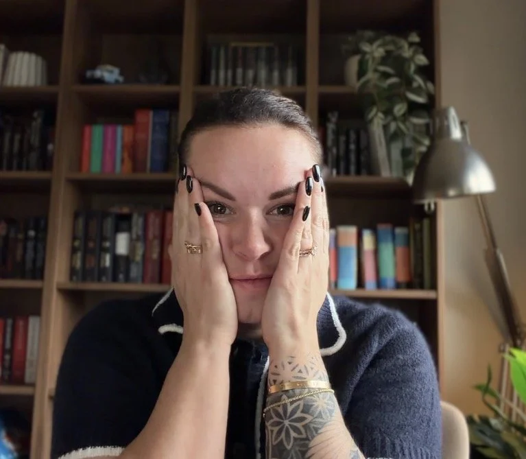 Woman with dark hair pulled back, wearing a dark sweater, holding her face with both hands, sitting in front of a bookshelf filled with colorful books and a desk lamp.