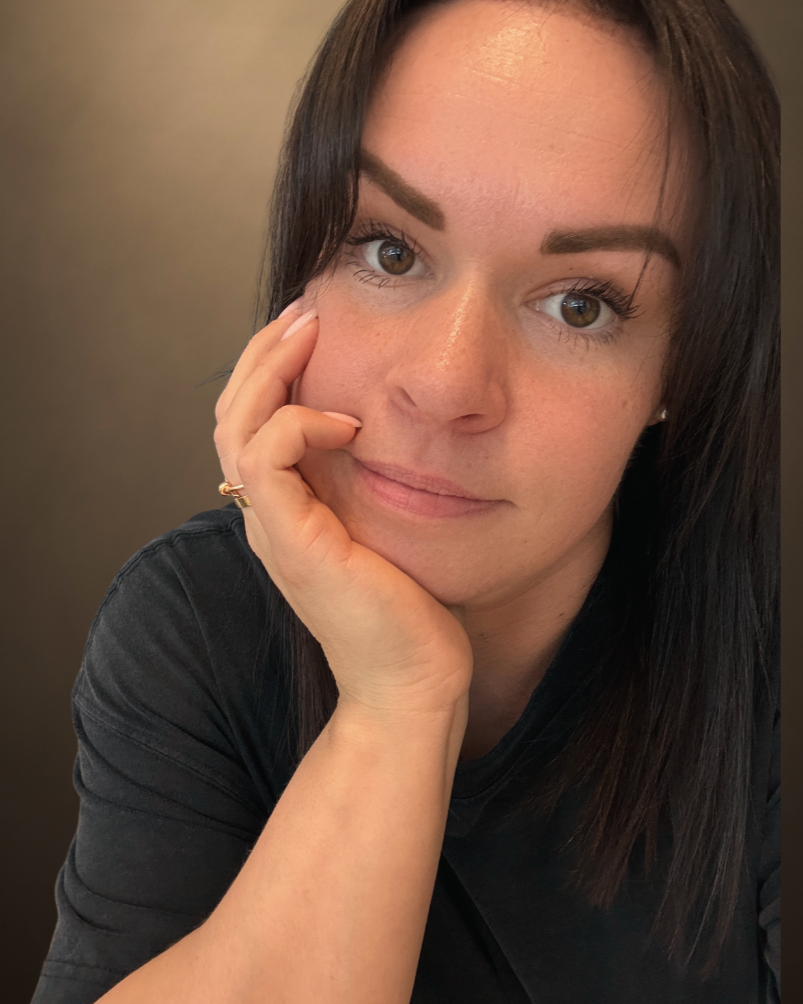 Close-up of a woman with dark brown hair, resting her face on her right hand, looking into the camera with a gentle smile.
