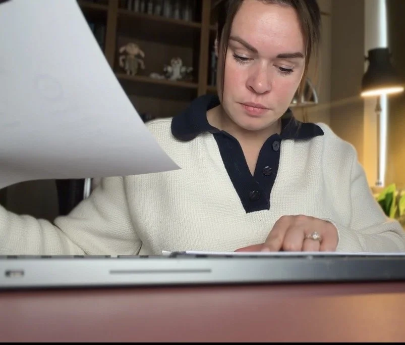 A woman working on a laptop at a desk, holding a piece of paper.