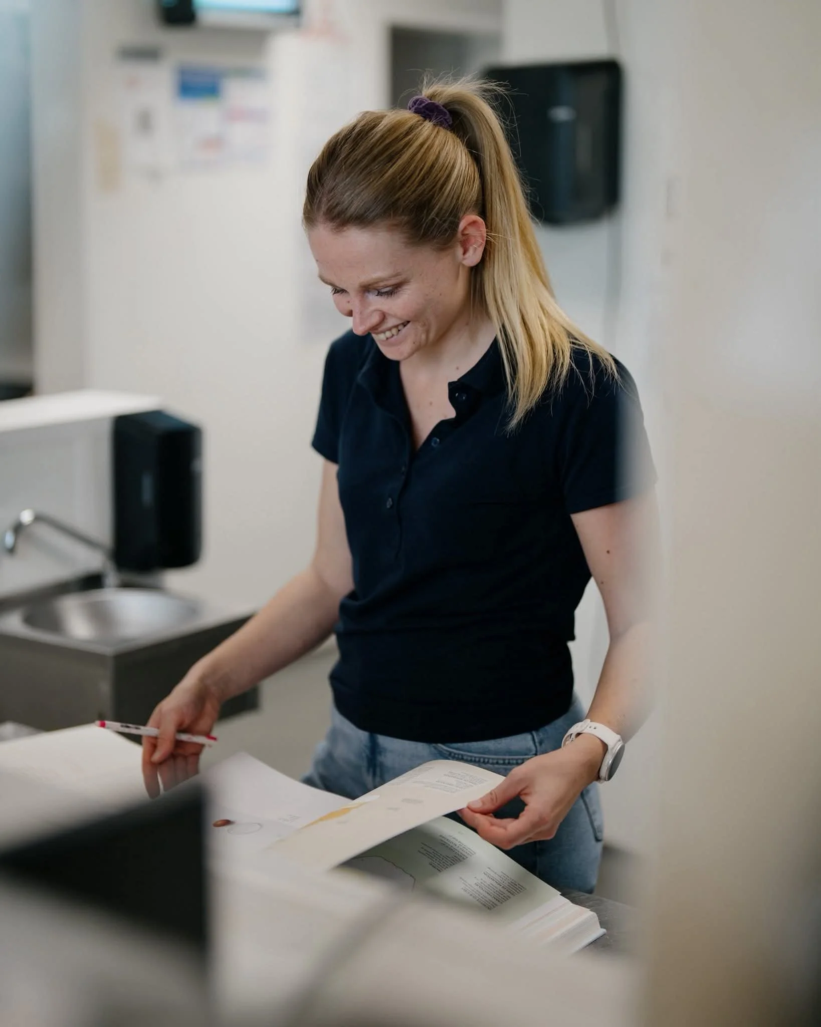 Une femme à cheveux blonds attachés en queue de cheval portant un polo noir, en train de lire ou de manipuler des documents ou des plans sur une table, dans un environnement intérieur, probablement un bureau ou un centre de travail.